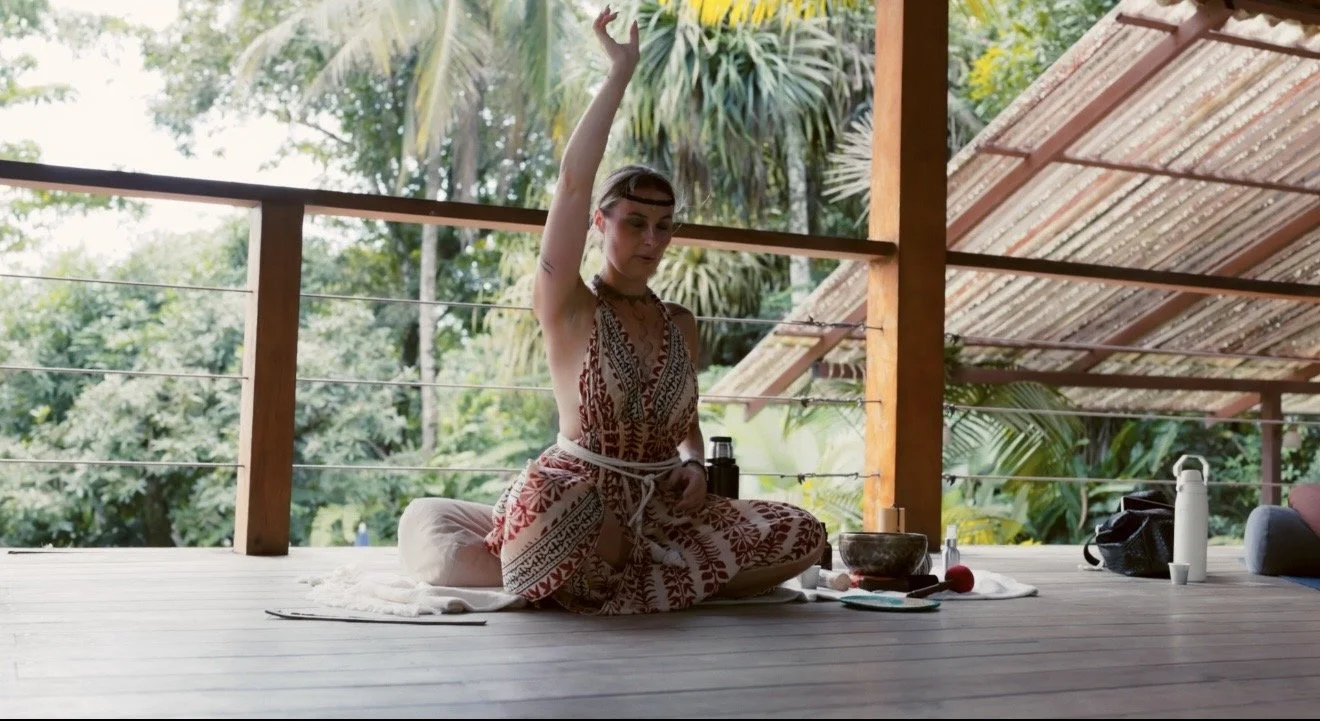 Young woman sitting cross-legged on a wooden porch, meditating with one arm raised, surrounded by greenery, with a prayer bowl and water bottle nearby.