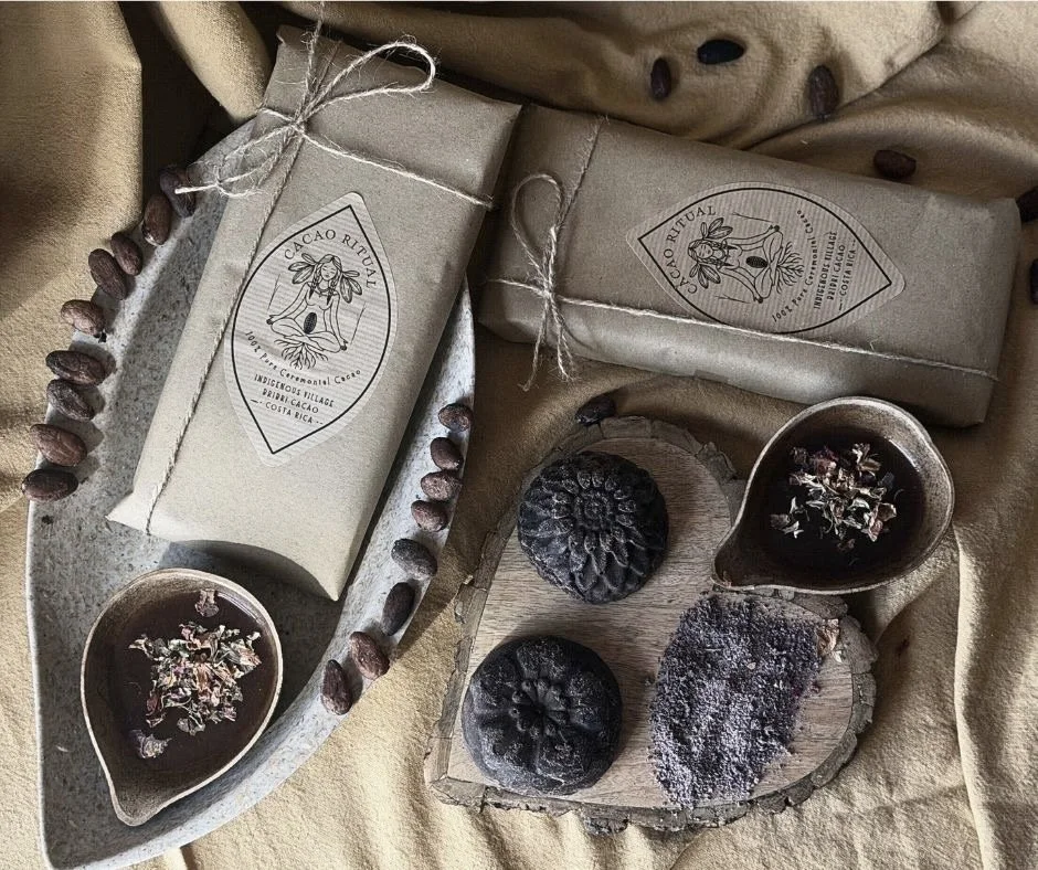 Two wrapped chocolate bars, two cups of hot chocolate topped with dried flower petals, two black cacao pods, and a piece of cacao nibs on a piece of wood, all arranged on a beige cloth with cacao beans scattered around.