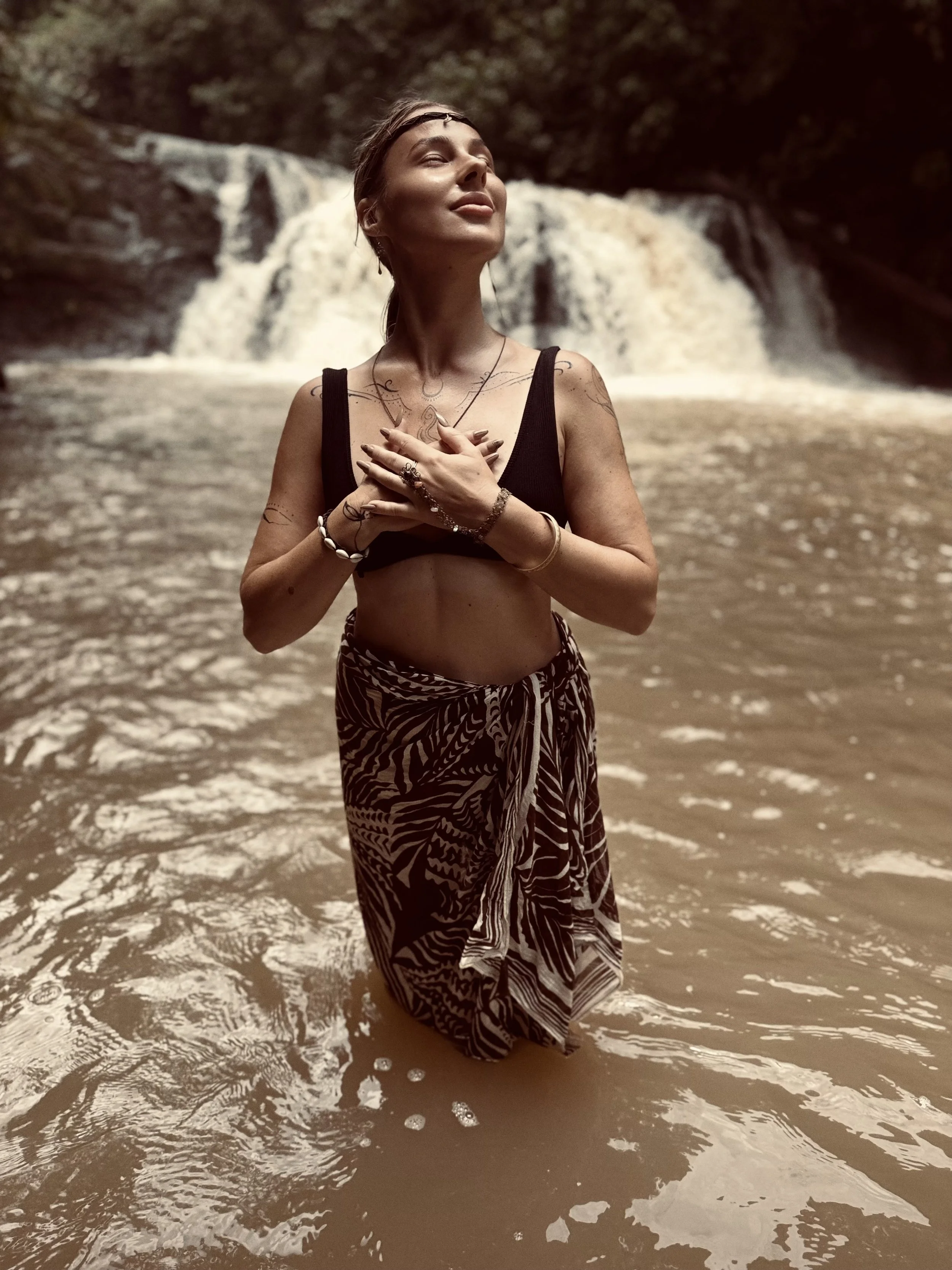 A woman standing in shallow water at the beach with her hands over her heart and eyes closed, smiling, with a waterfall and trees in the background.