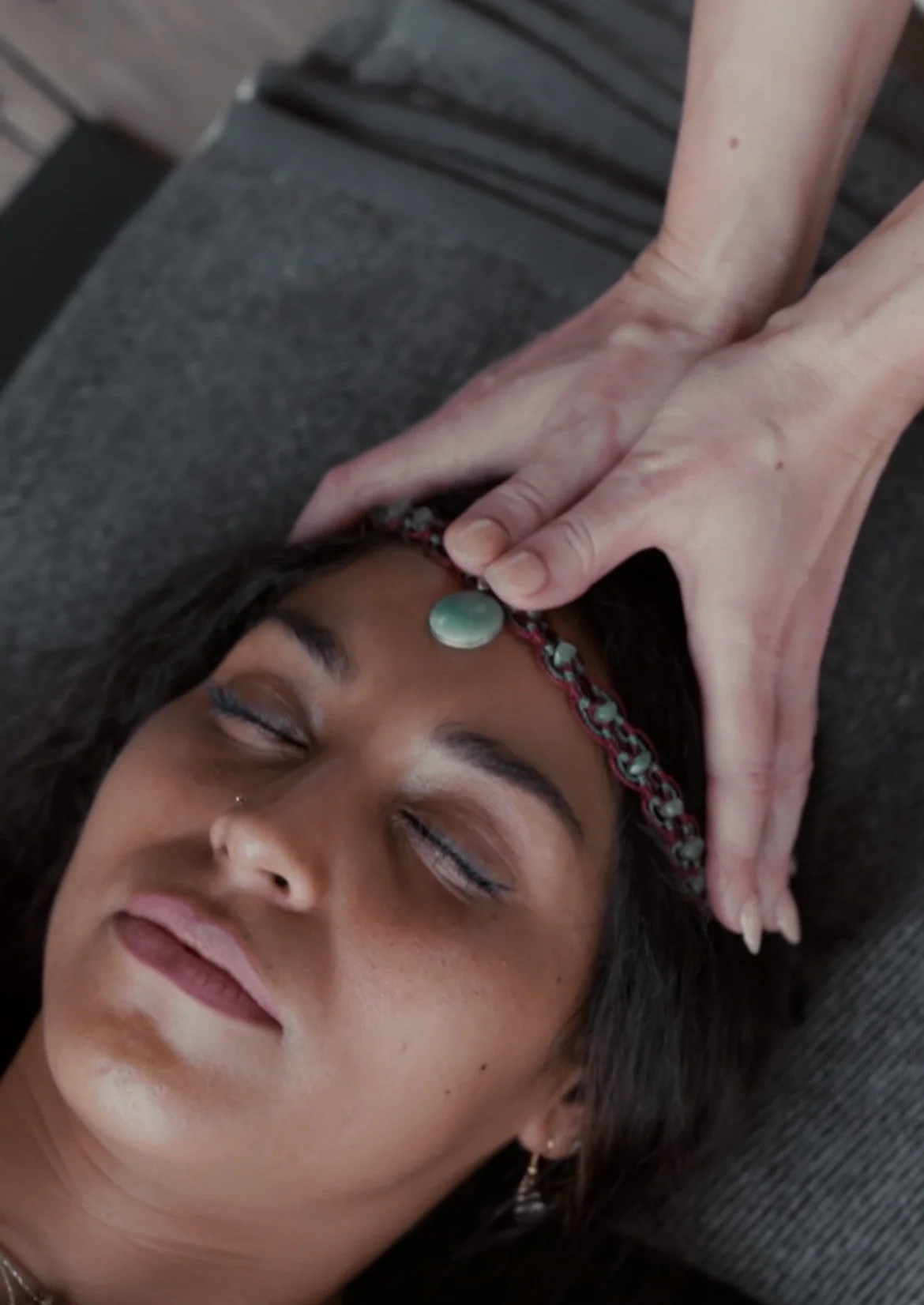 A woman receiving a head massage, lying down with eyes closed, wearing a colorful headband and earrings.