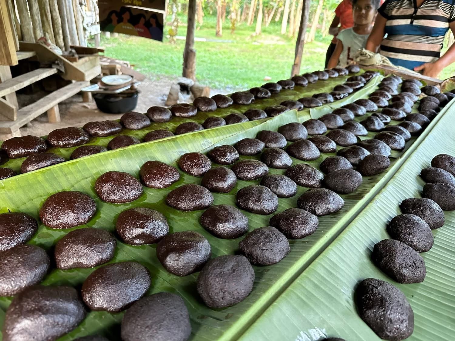 Rows of small, round, dark brown or black balls, possibly food items, placed on long banana leaves on a table outdoors.