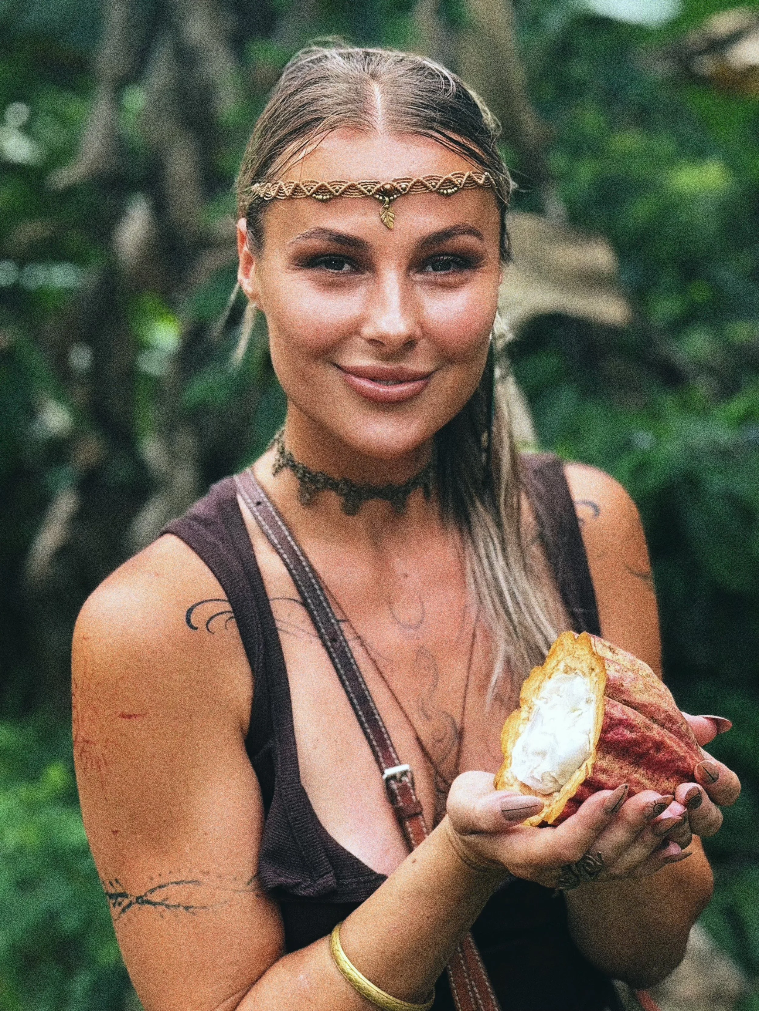 A woman with braided hair, wearing bohemian jewelry and a sleeveless top, holding a pineapple with a generous amount of whipped cream inside, standing outdoors with green foliage in the background.
