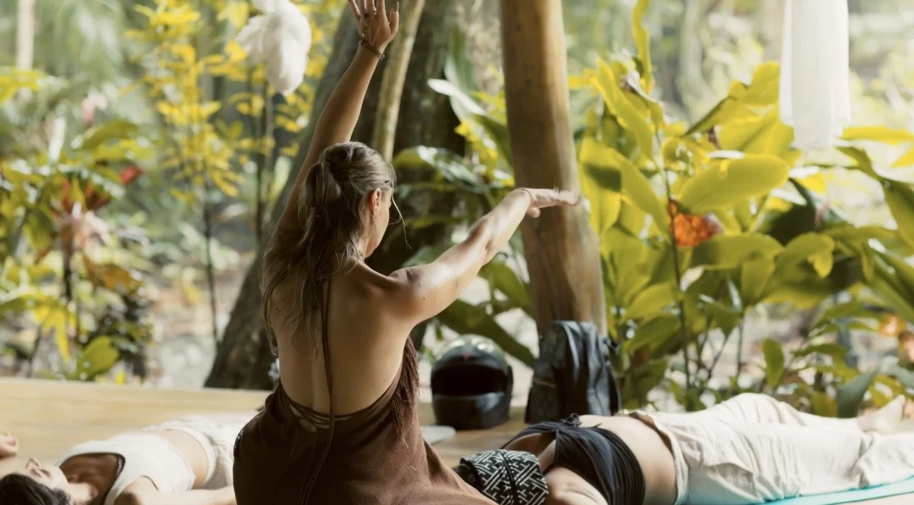 A woman leading a yoga class outdoors, with people lying on yoga mats in a forest setting, practicing meditation or relaxation exercises.