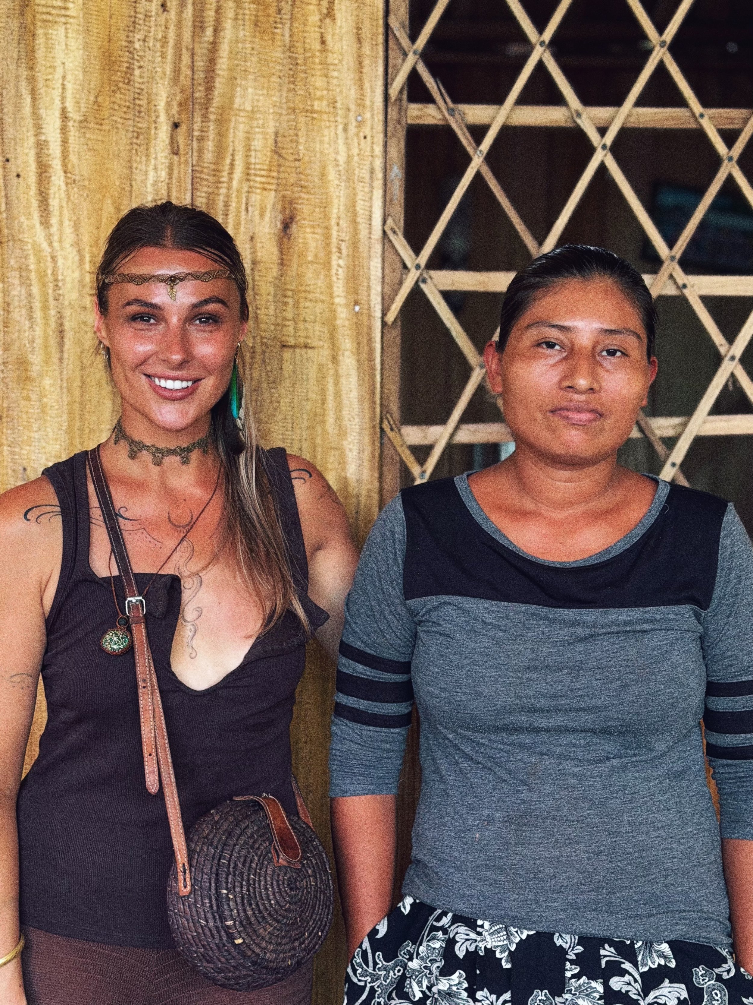 A woman with long hair and tattoos on her arm standing next to a young man with short hair, both smiling and standing in front of wooden and lattice background.