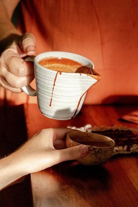 A person holding a ceramic mug with coffee or hot chocolate and red syrup dripping down the outside, with another hand holding a small wooden bowl on a wooden table.