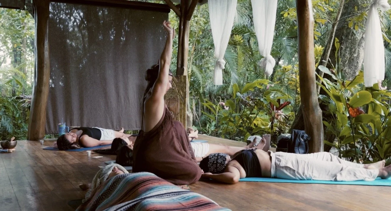 People practicing yoga outdoors on mats under a wooden canopy, surrounded by lush green foliage.