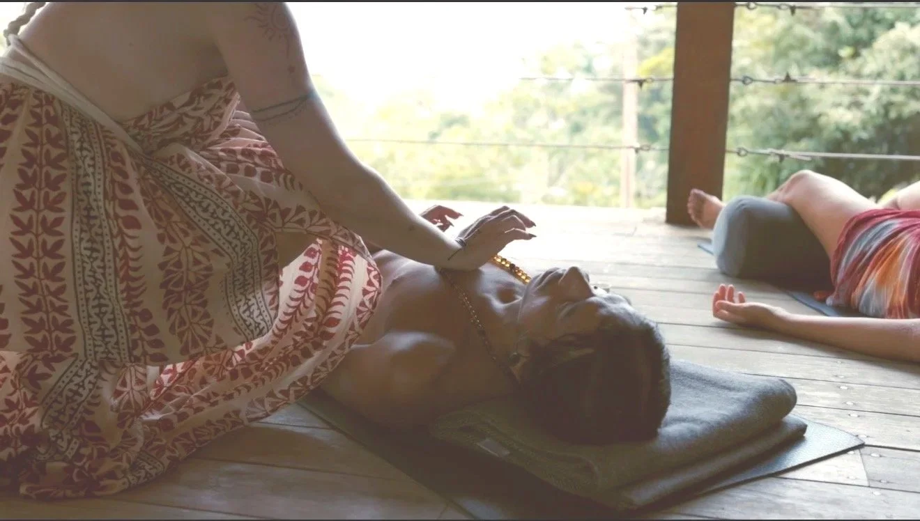 Two people practicing yoga outdoors on a wooden deck surrounded by trees. One person is lying on their back with legs bent and arms relaxed, and the other person is sitting cross-legged, gently touching the first person's stomach.