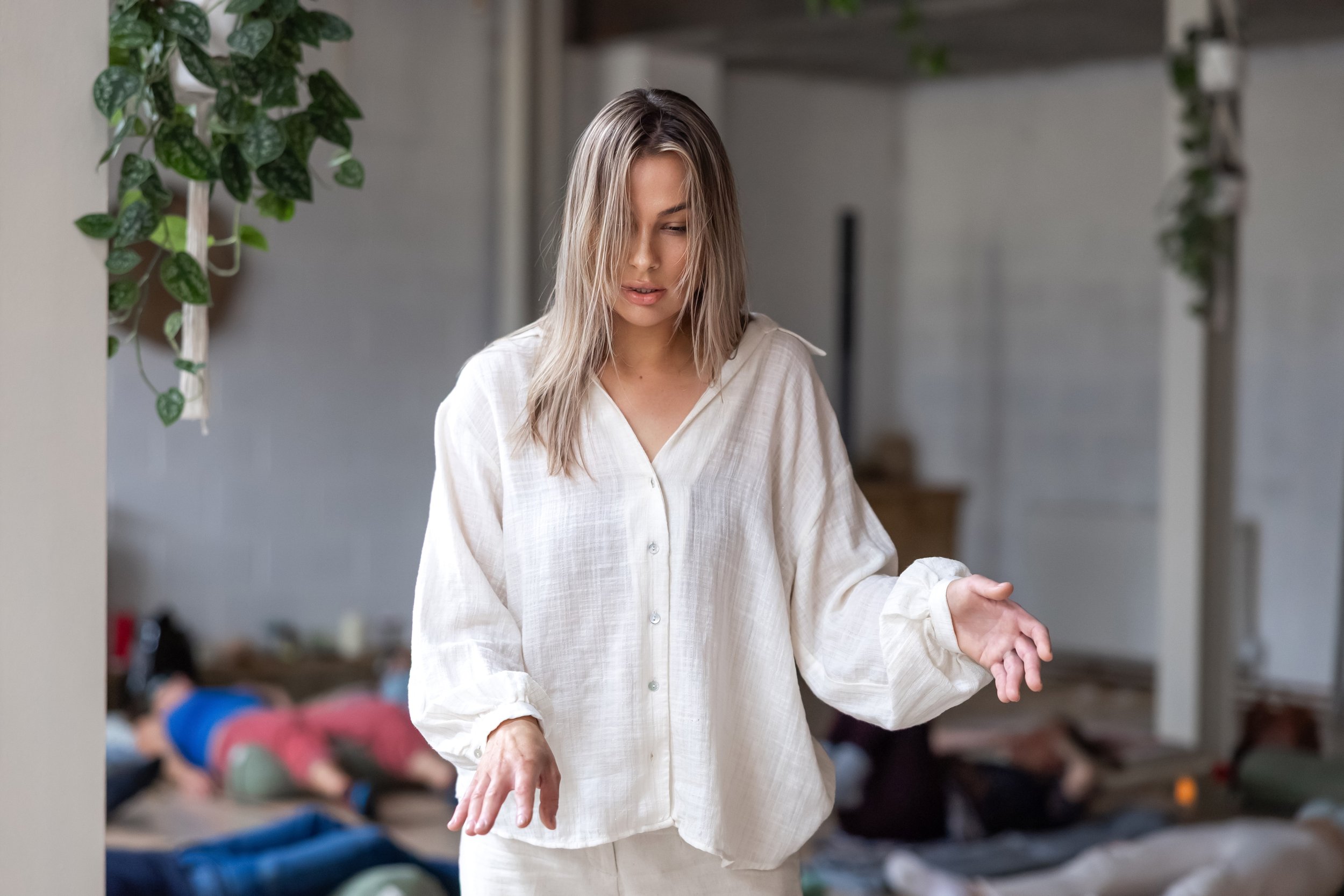 A woman with light brown hair stands in a room, looking down with a thoughtful expression. She wears a loose, white shirt and appears to be in a calm, contemplative state. In the background, there are multiple people lying on their stomachs on the floor, and the room is decorated with green plants.