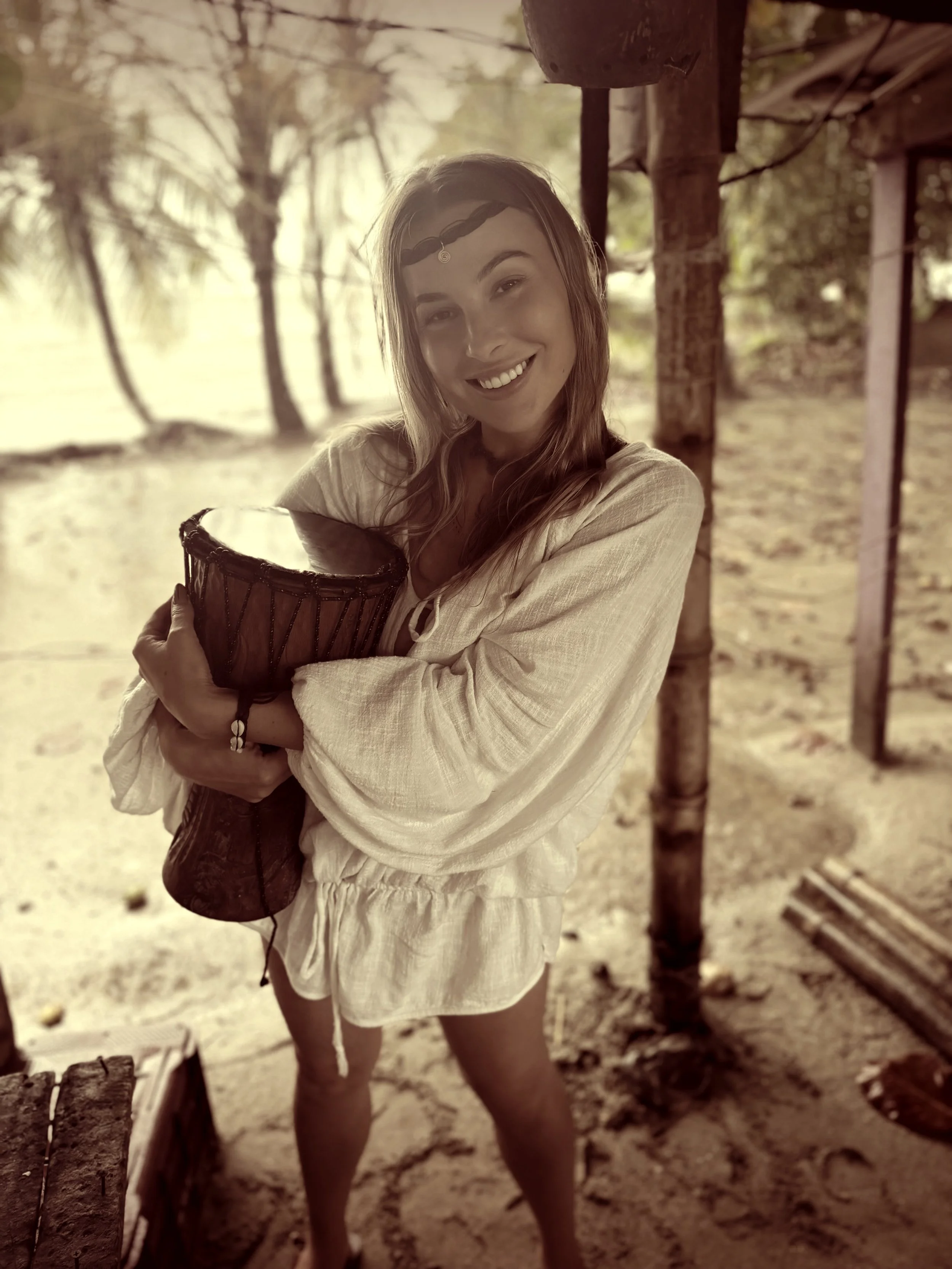 A young woman with long hair wearing a headband and white dress, smiling and holding a drum, standing outdoors with trees and a wooden structure in the background.