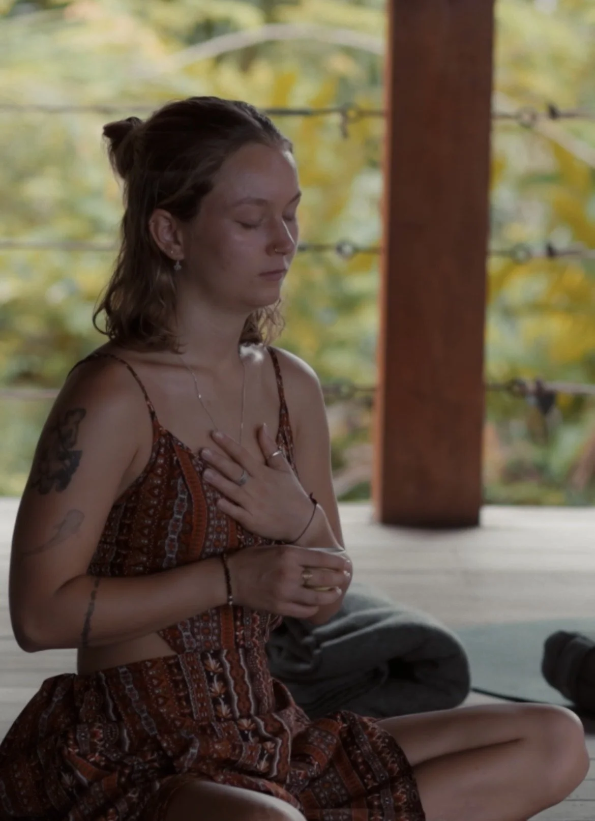 A young woman with tattoos and jewelry is sitting cross-legged outdoors, with her eyes closed and her hand on her chest, appearing to be meditating or in deep thought.