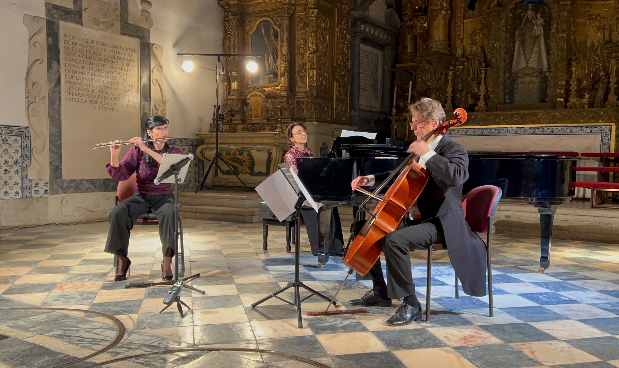 Três músicos tocando instrumentos em um palco, incluindo uma flautista, uma pianista e um violoncelista, em um ambiente de igreja ou teatro antigo com decoração dourada e piso de ladrilhos.