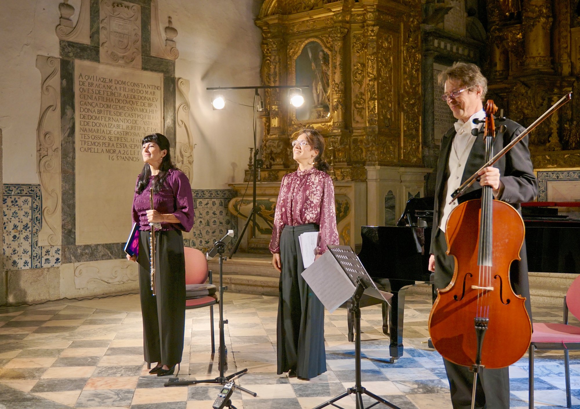 Três músicos em uma apresentação em um interior de igreja com decoração dourada e uma parede com inscrição em português, incluindo uma mulher segurando uma flauta, uma mulher de pé com papel na mão, e um homem com violoncelo, todos de pé em frente ao