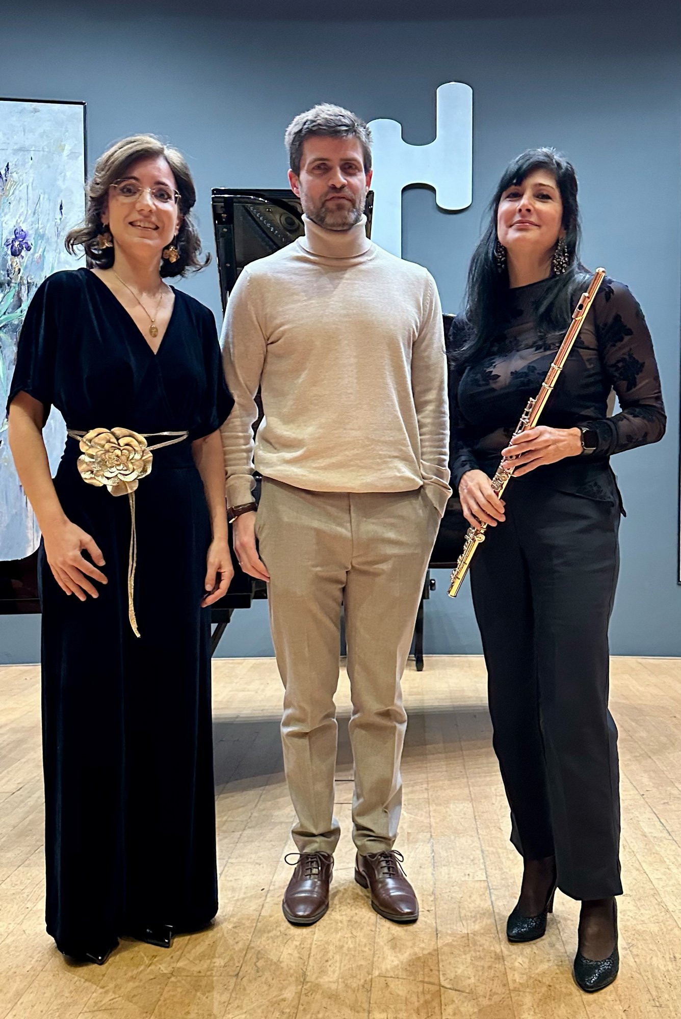 Três músicos posando em um palco, uma mulher com vestido preto, uma homem de terno bege, e outra mulher com blusa de tule preta e segurando uma flauta.