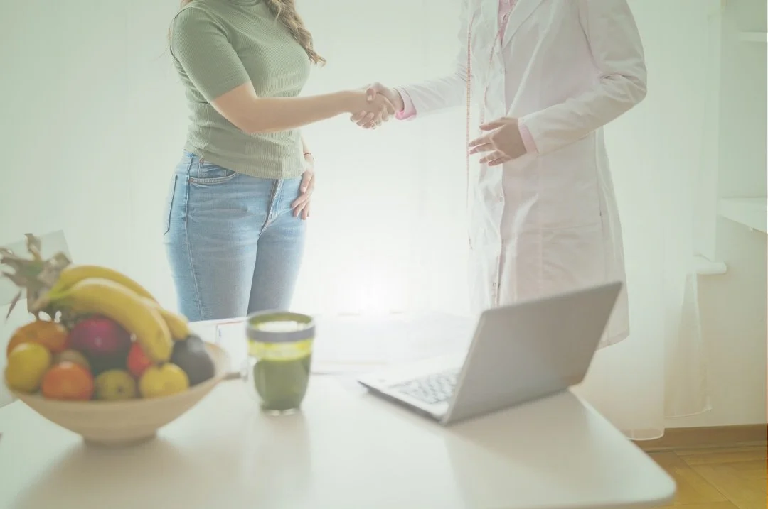 Una donna e un medico si stringono la mano in un ambiente luminoso, con una frutta su un tavolo e un laptop acceso.