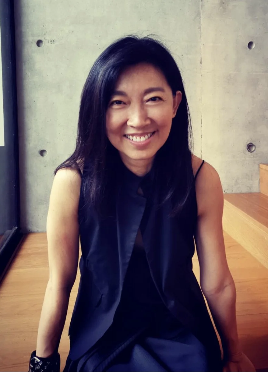 A woman with black hair and a black sleeveless top, smiling, sitting on a wooden surface in front of a concrete wall.