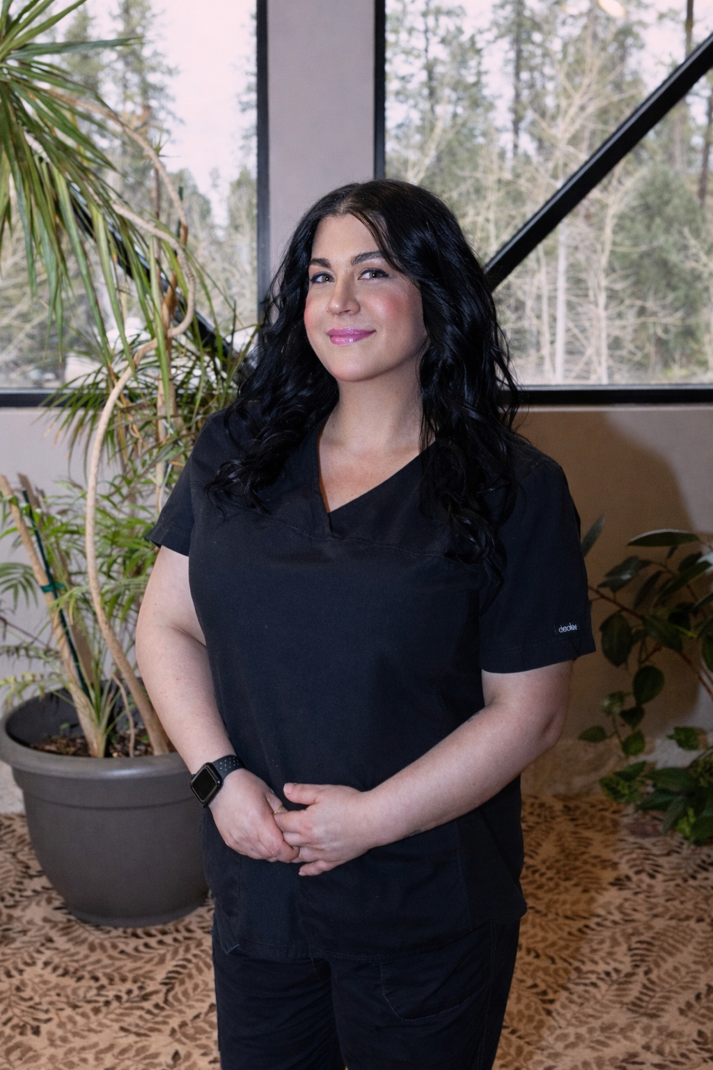 A woman with long black hair wearing black scrubs and a smartwatch, standing indoors in front of large windows with trees outside, surrounded by houseplants on a leopard print rug.