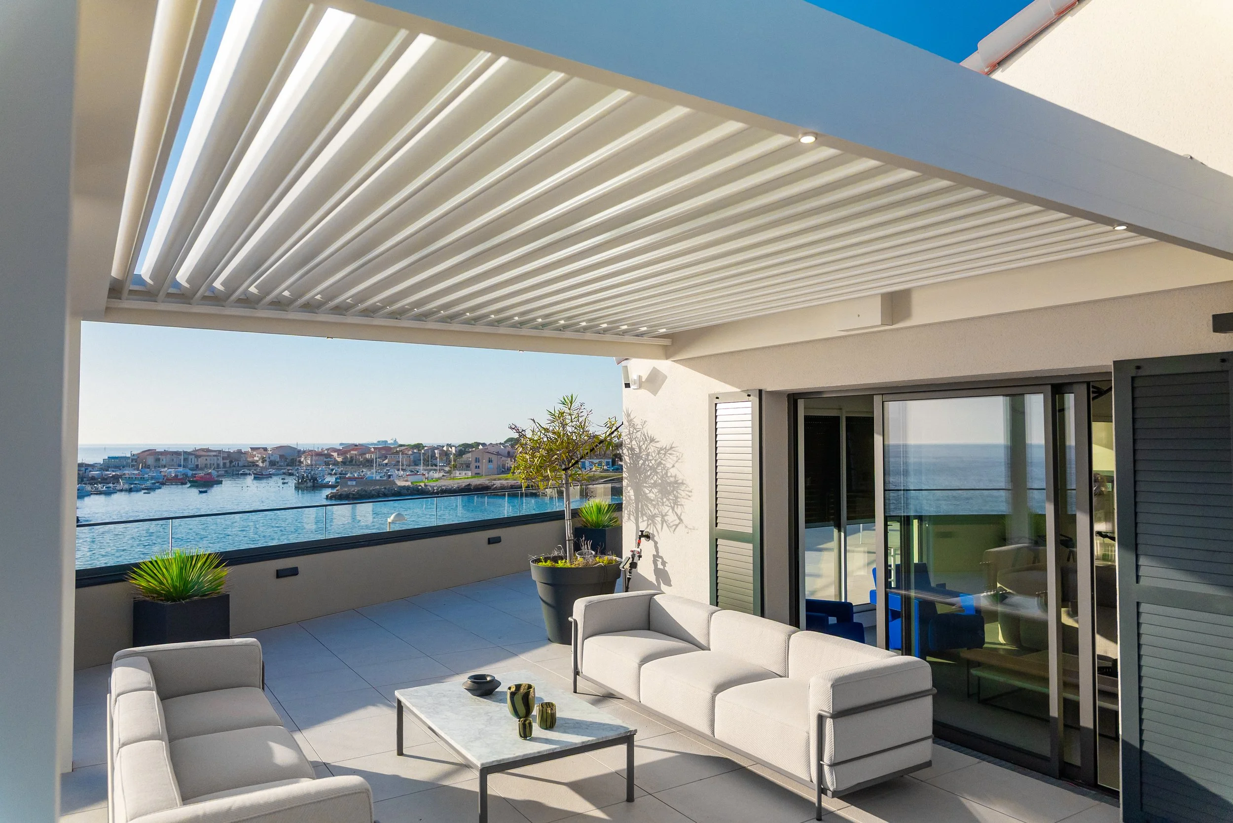 Terrasse moderne avec vue sur la mer, meubles d'extérieur, plantes et pergola en métal blanc.