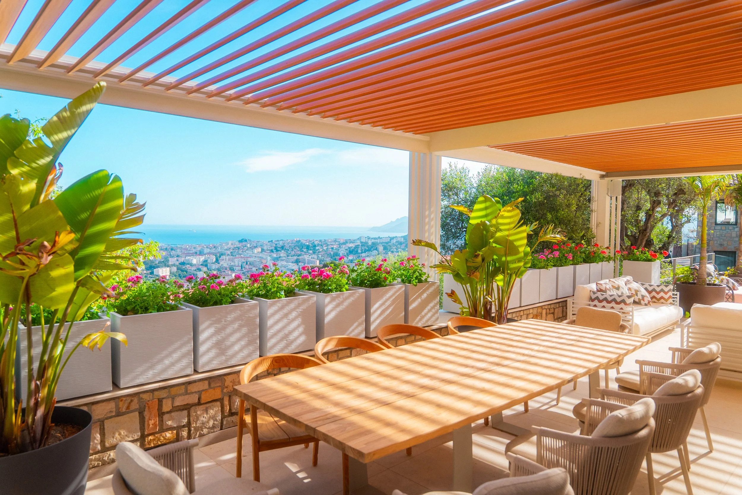 Terrasse ensoleillée avec vue sur la ville, plantes vertes, table en bois et fauteuils confortables.