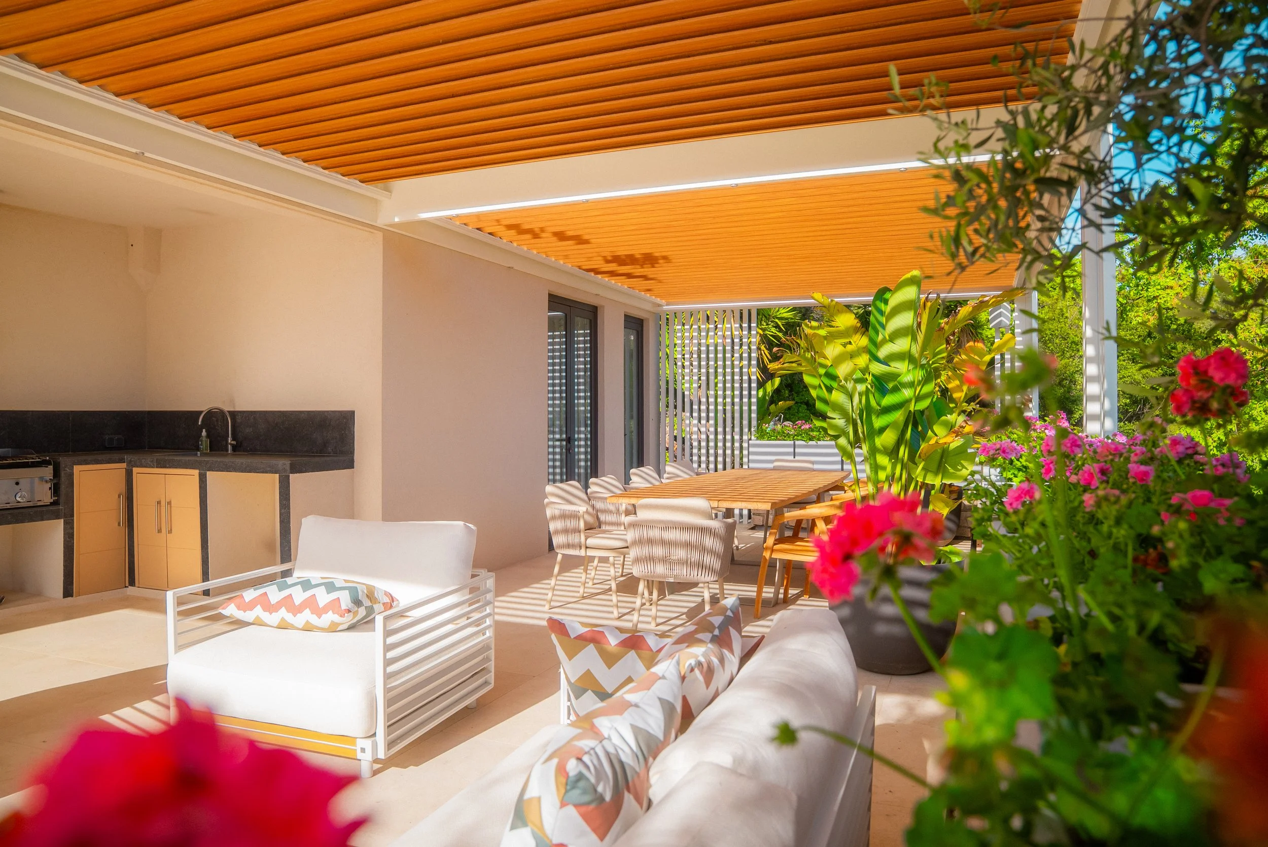 Terrasse extérieure avec mobilier de jardin, plantes vertes et fleurs, plafond en bois, espace repas avec grande table et chaises, coin détente avec fauteuils et canapé.