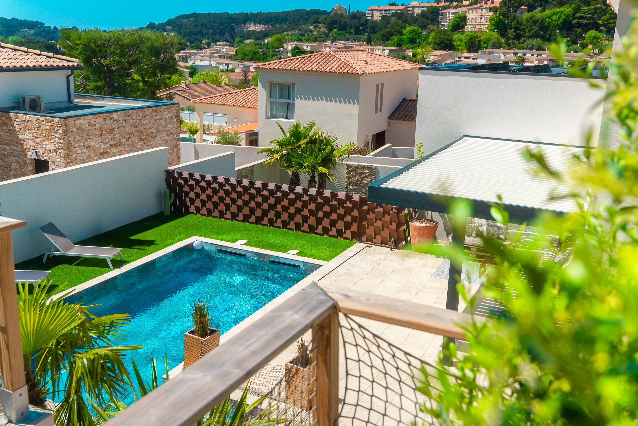 Vue d'une piscine extérieure dans un jardin privé avec détenteur, plantes tropicales, murs de clôture, maisons environnantes et collines en arrière-plan sous un ciel bleu clair.