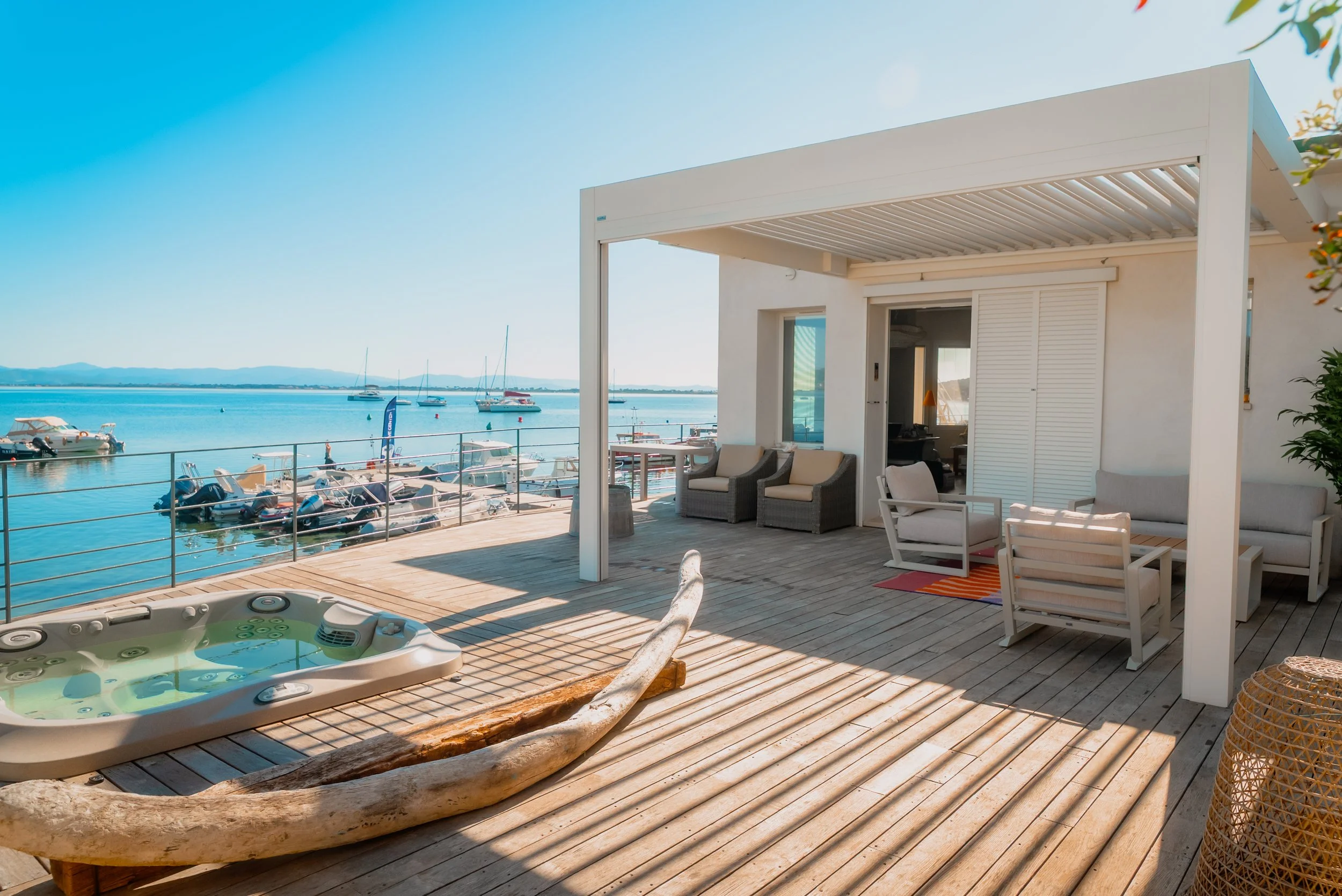 Terrasse en bois avec jacuzzi, meubles confortables, vue sur un port avec bateaux, ciel bleu clair, ambiance relaxante.