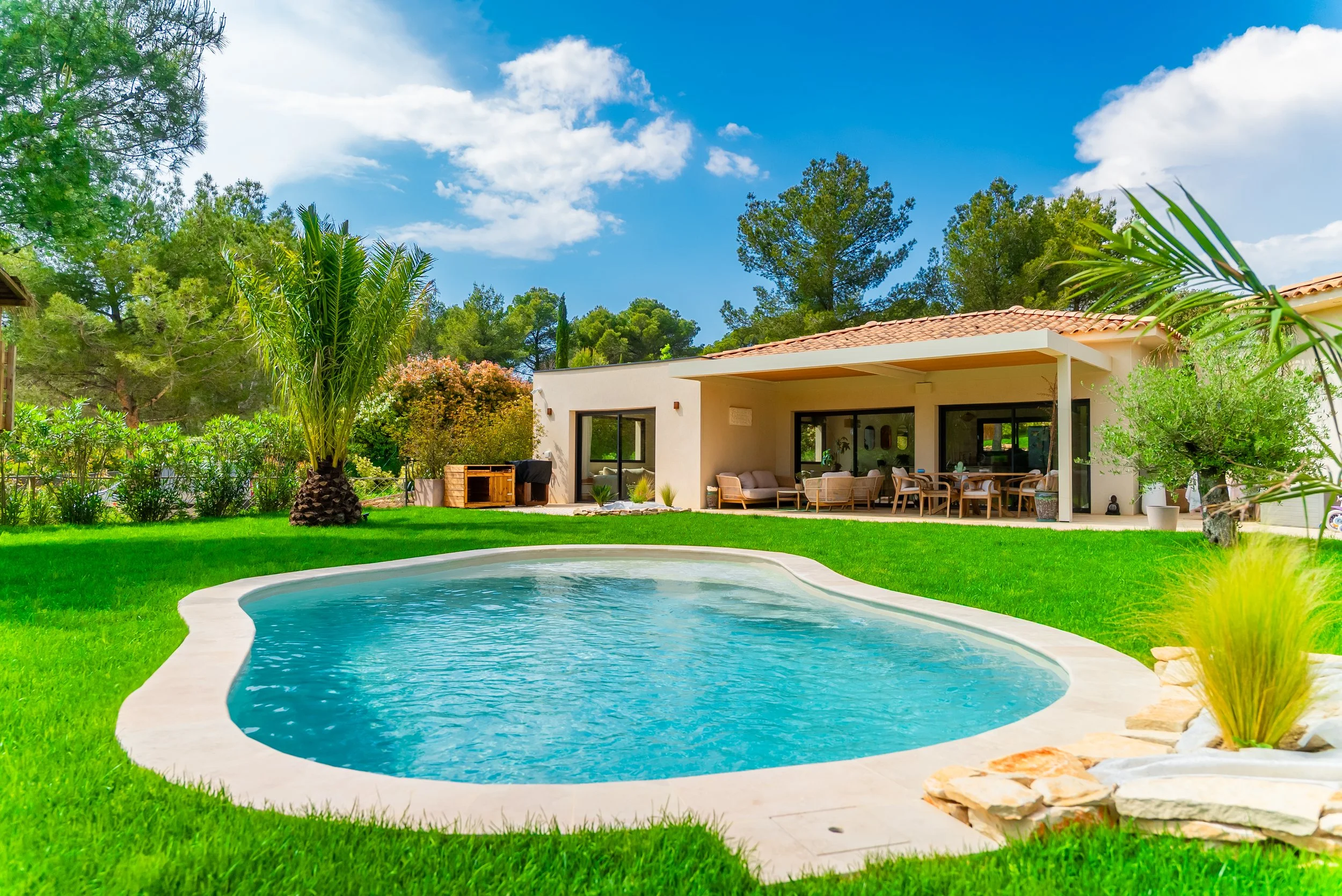 Maison moderne avec terrasse et piscine dans un jardin verdoyant, arbres et ciel bleu avec quelques nuages.
