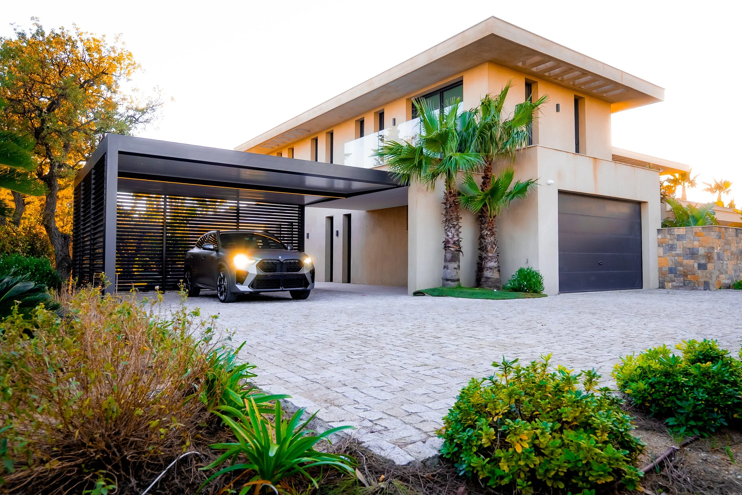 Vue d'une maison moderne avec un garage, des palmiers et un véhicule noir garé sous une structure de carport en métal noir, pelouse et végétation environnante.