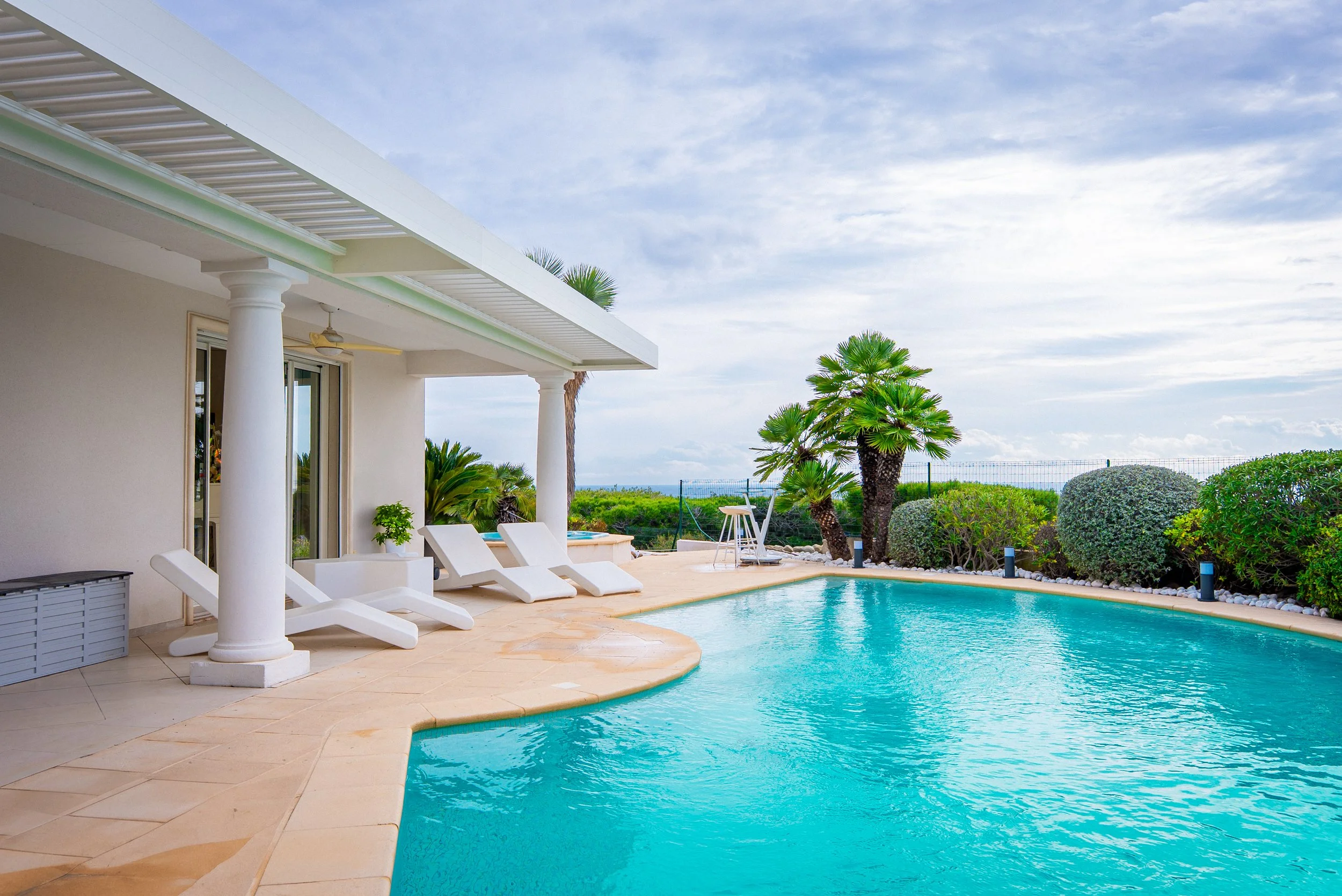 Piscine extérieure avec chaises longues, abri avec colonnes, végétation tropicale et vue sur le ciel.