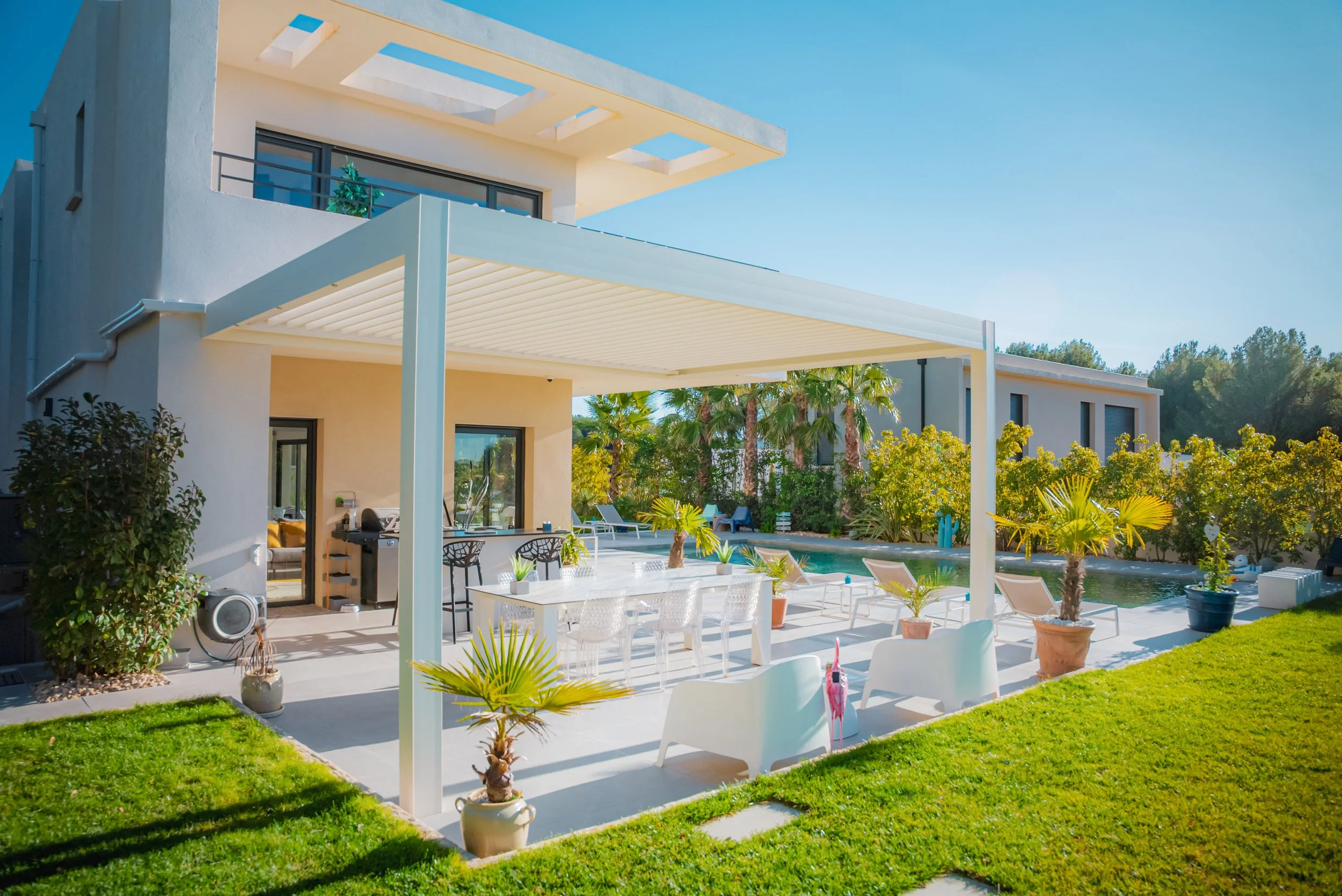 Une maison moderne avec un patio et une piscine entourée de plantes tropicales, sous un ciel bleu clair.