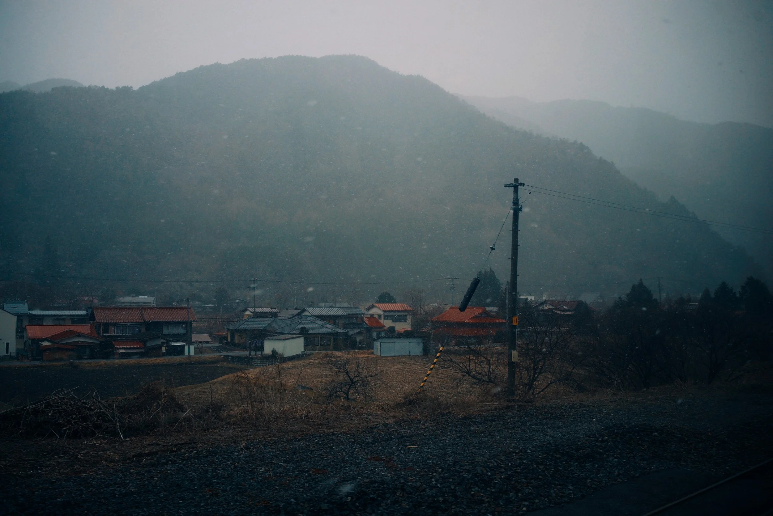 A rural landscape with small houses, a utility pole with leaning wires, and mountains in the background, under overcast skies with light snowfall.