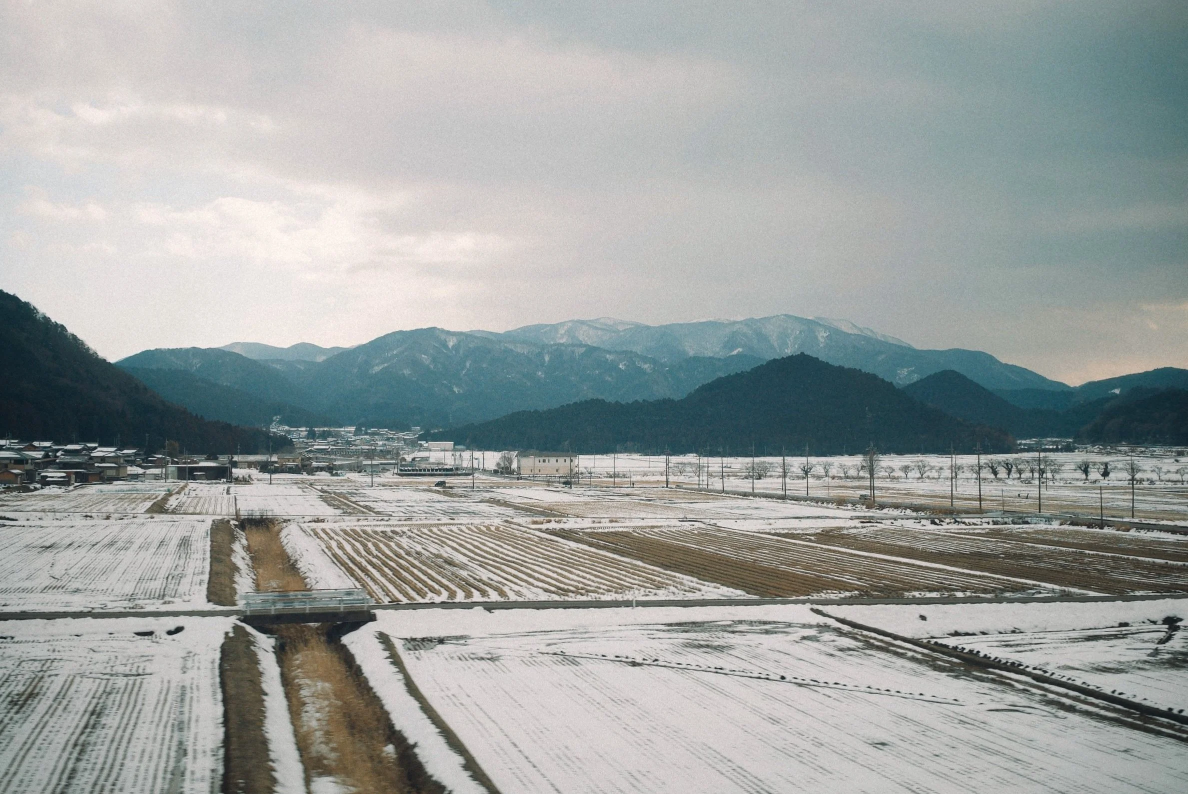 Snow-covered agricultural fields with mountains in the background, cloudy sky, and sparse trees.