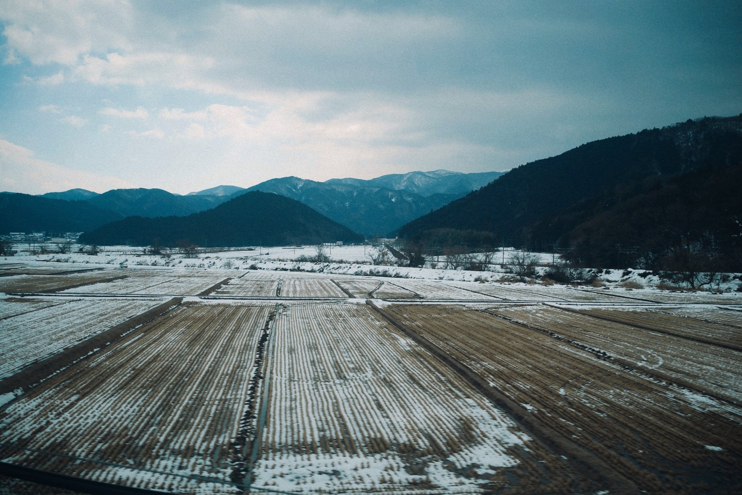 Snow-covered farmland with mountains in the background under a cloudy sky.