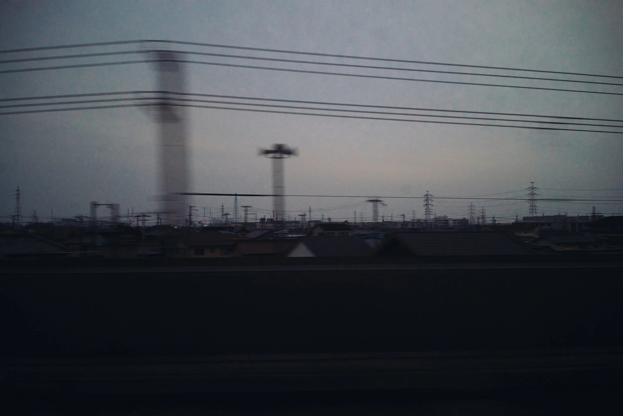 View of a city skyline with silhouette rooftops and tall utility poles, seen through a blurred window or glass, during dusk or dawn.