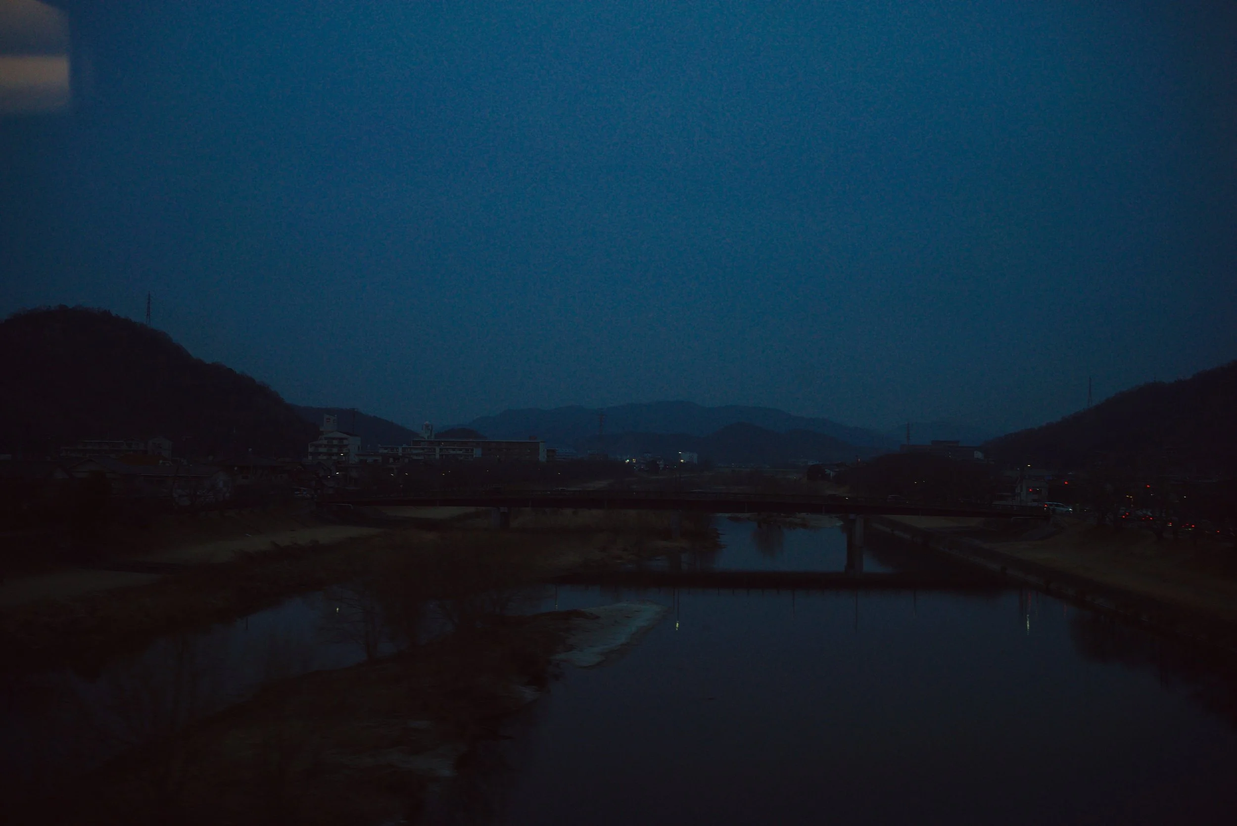 Nighttime landscape with a river, bridge, and distant hills under a dark blue sky.