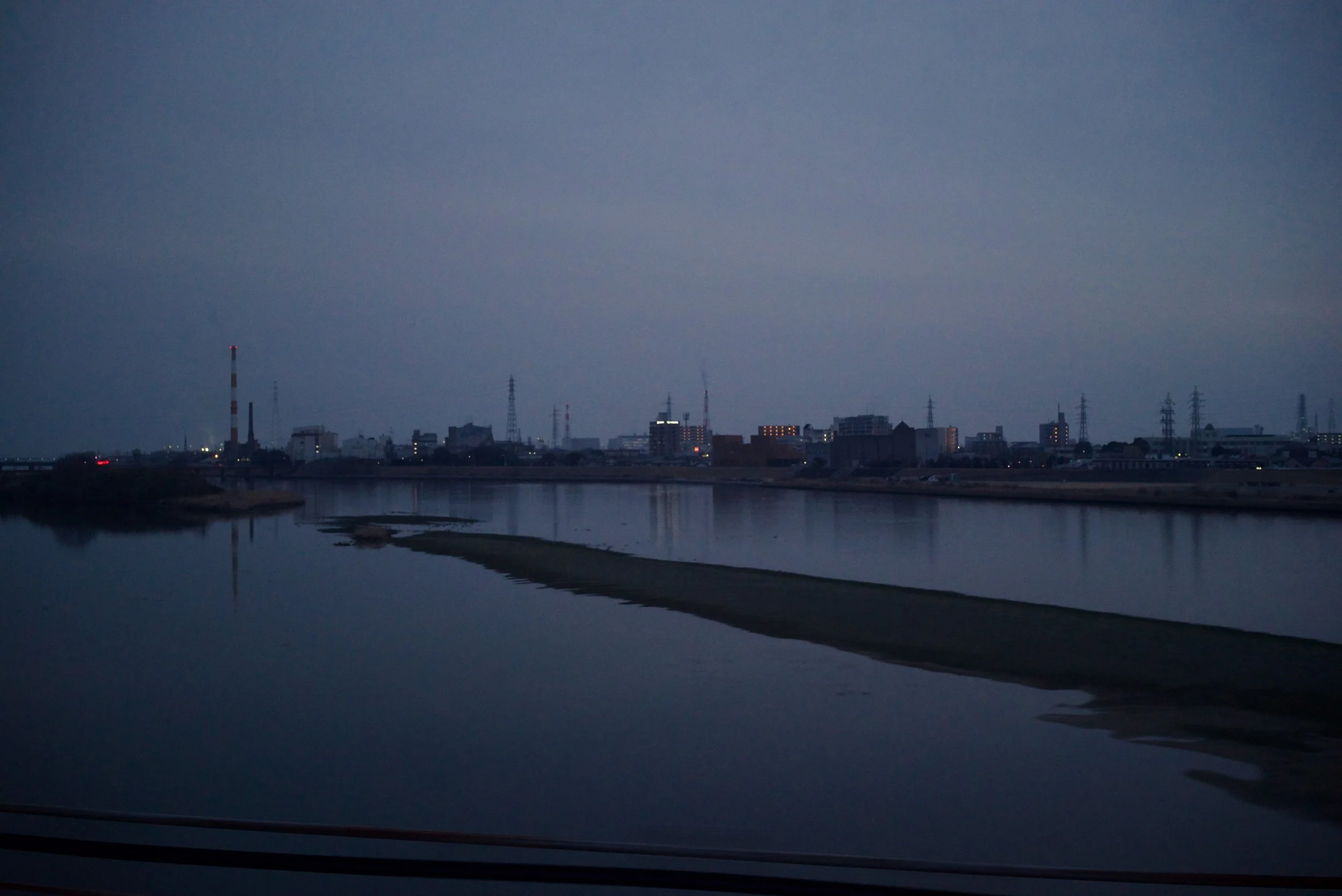 Cityscape at dusk with a river in the foreground and buildings with lights in the background.