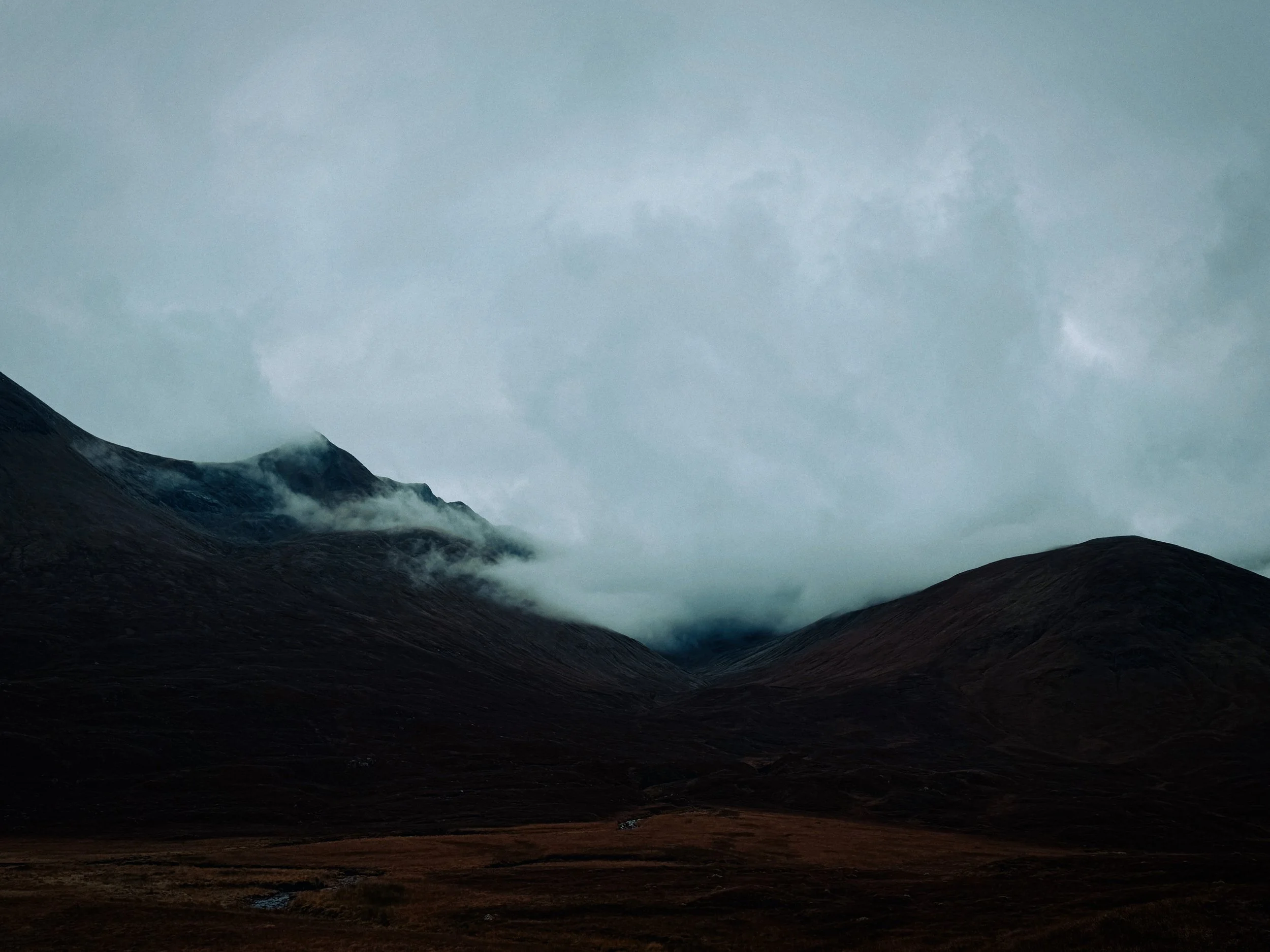 A dark, cloudy mountain landscape with mist and low clouds covering the peaks and a wide open valley in foreground.