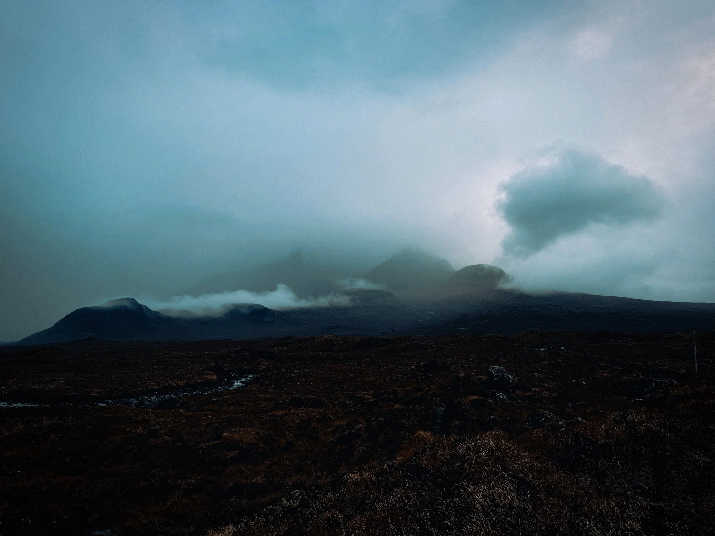 Mountain landscape with dark terrain, cloudy sky, and mist covering the mountain peaks.