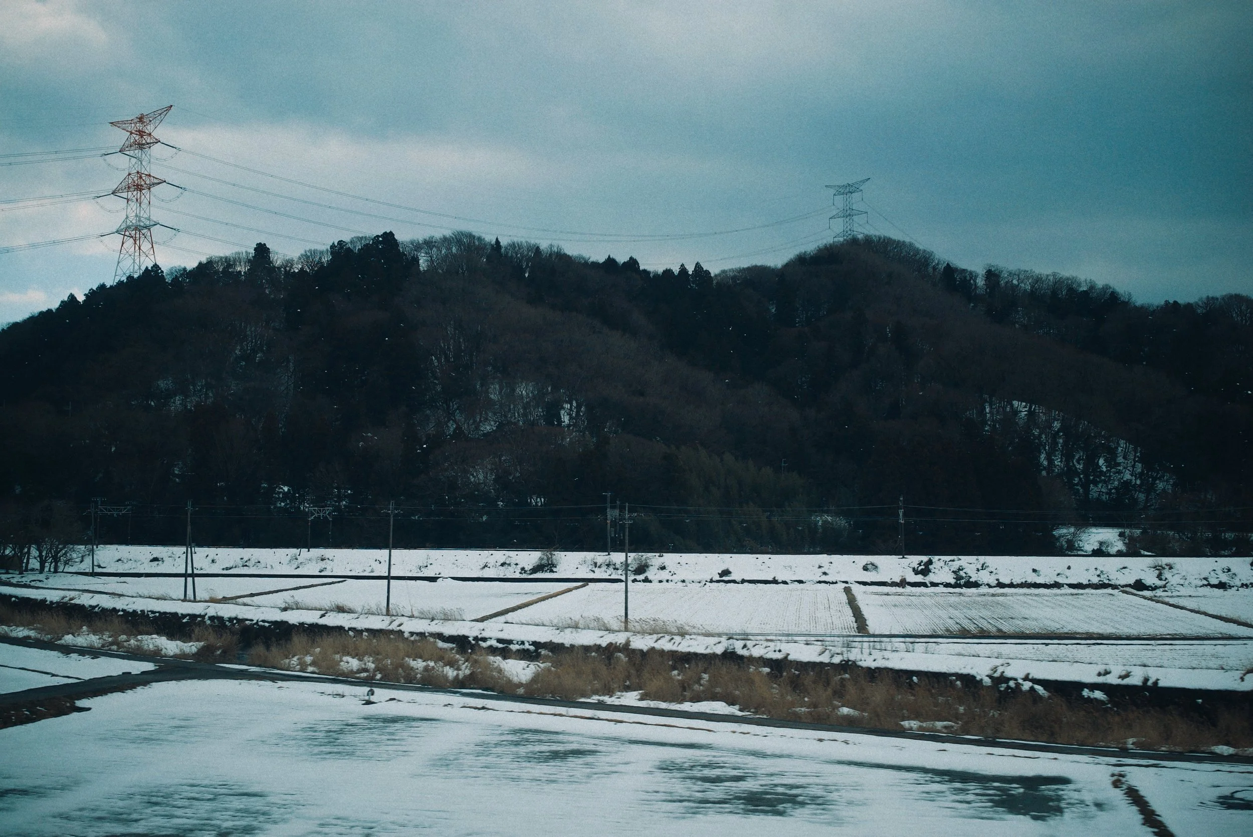 Snow-covered fields and a hill with trees, above which are power lines and electrical towers under a cloudy sky.