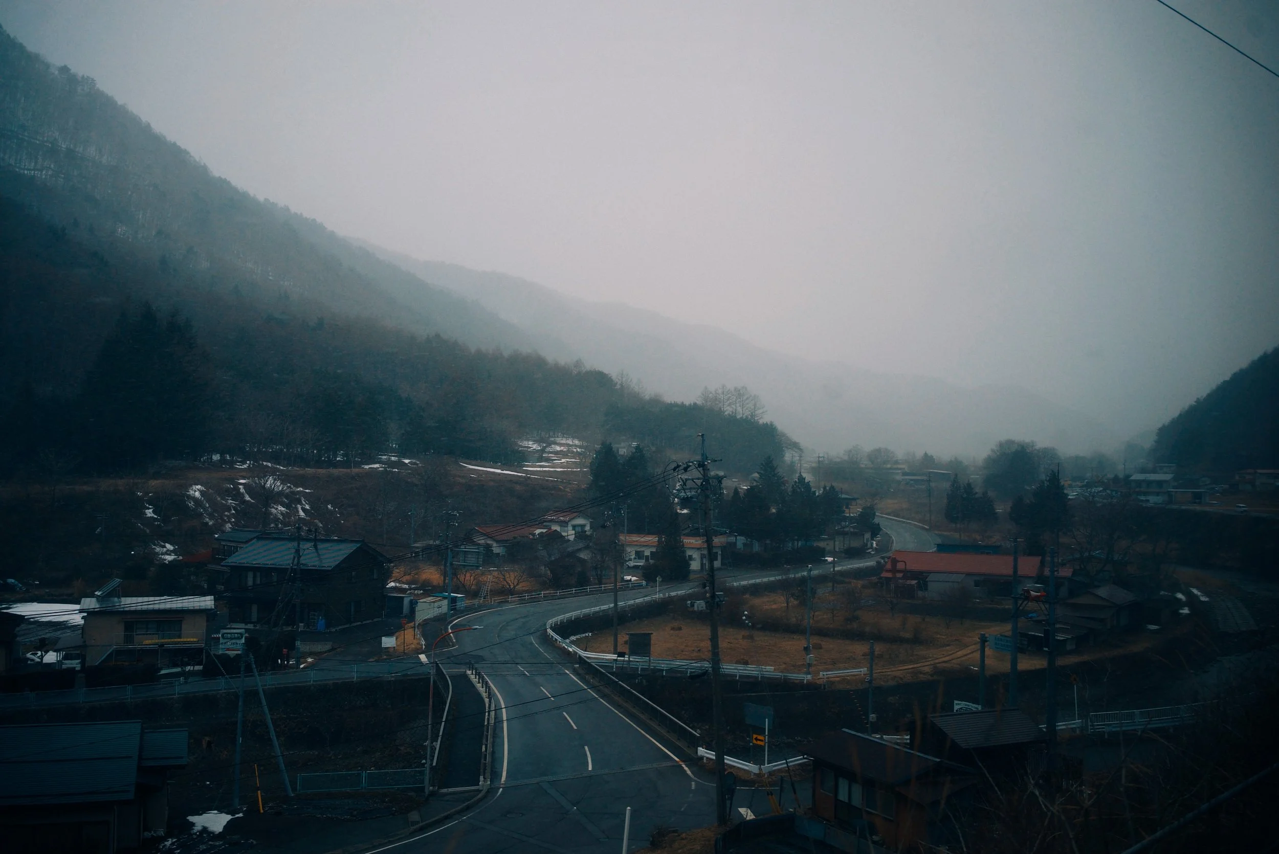 A foggy mountain village with winding road, sparse houses, and leafless trees.