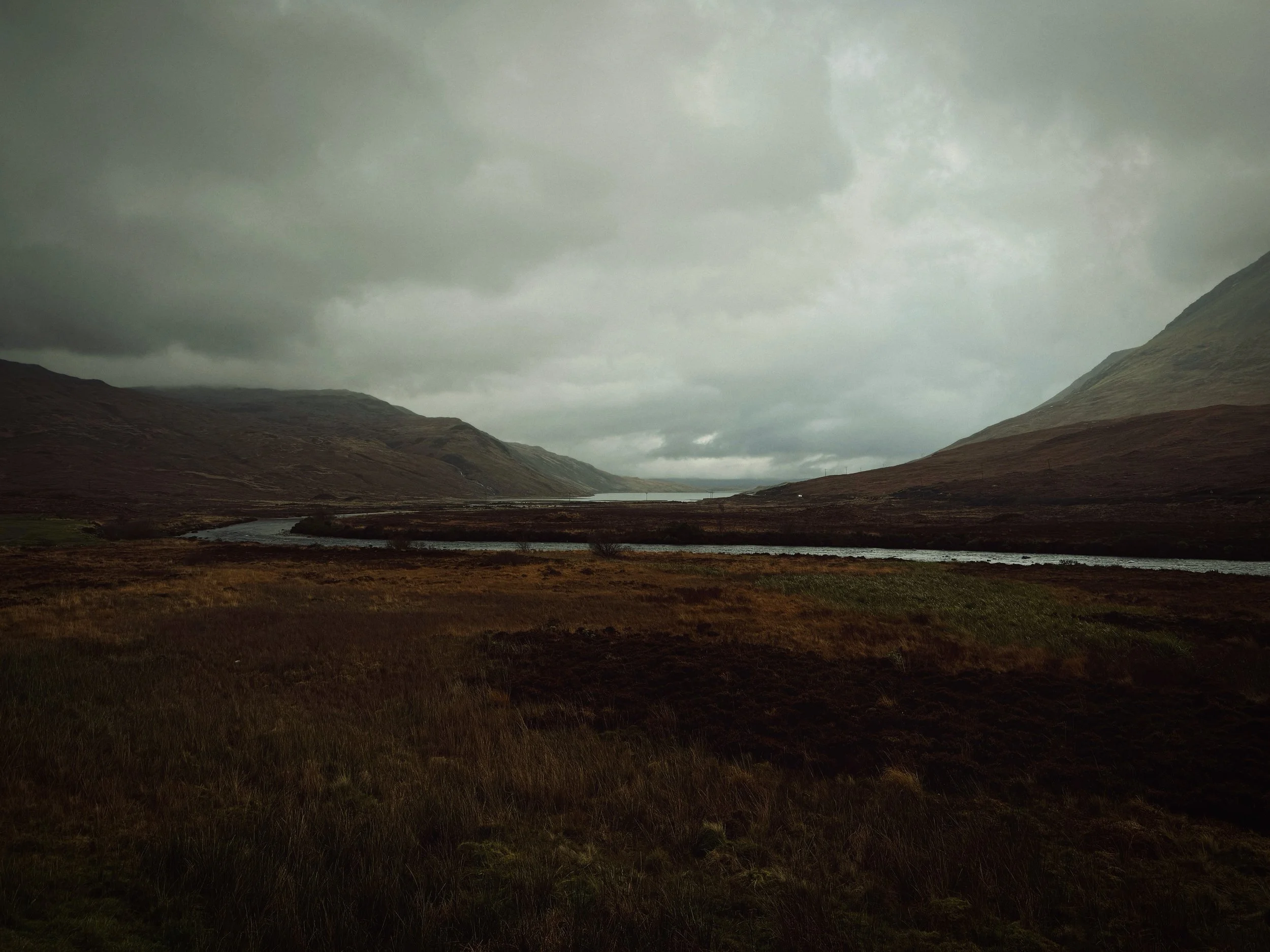 A landscape of a valley with dark mountains on either side, a river flowing through the center, and dark cloudy skies overhead.
