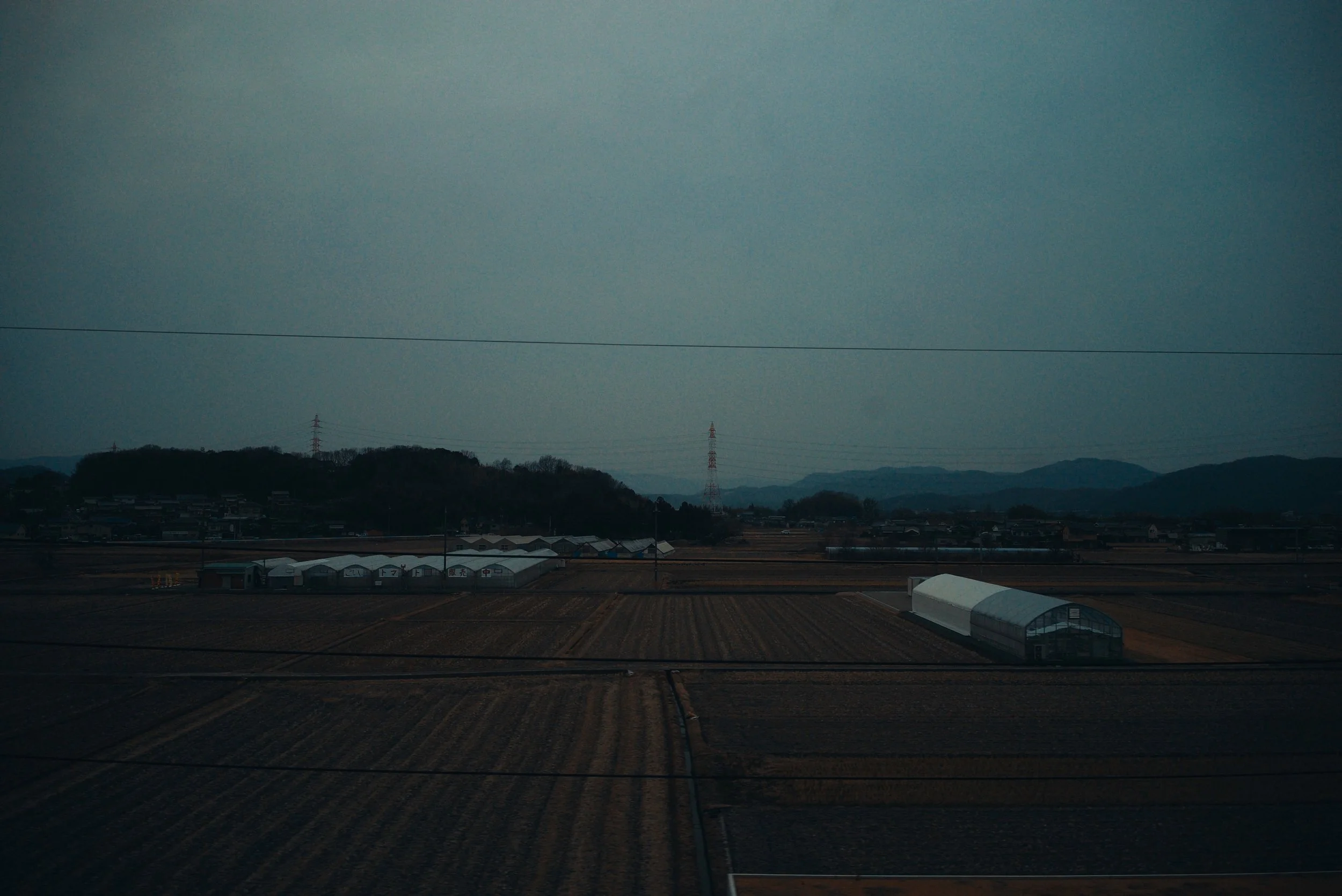 Agricultural fields with greenhouses on a cloudy day, distant mountains, and power lines.