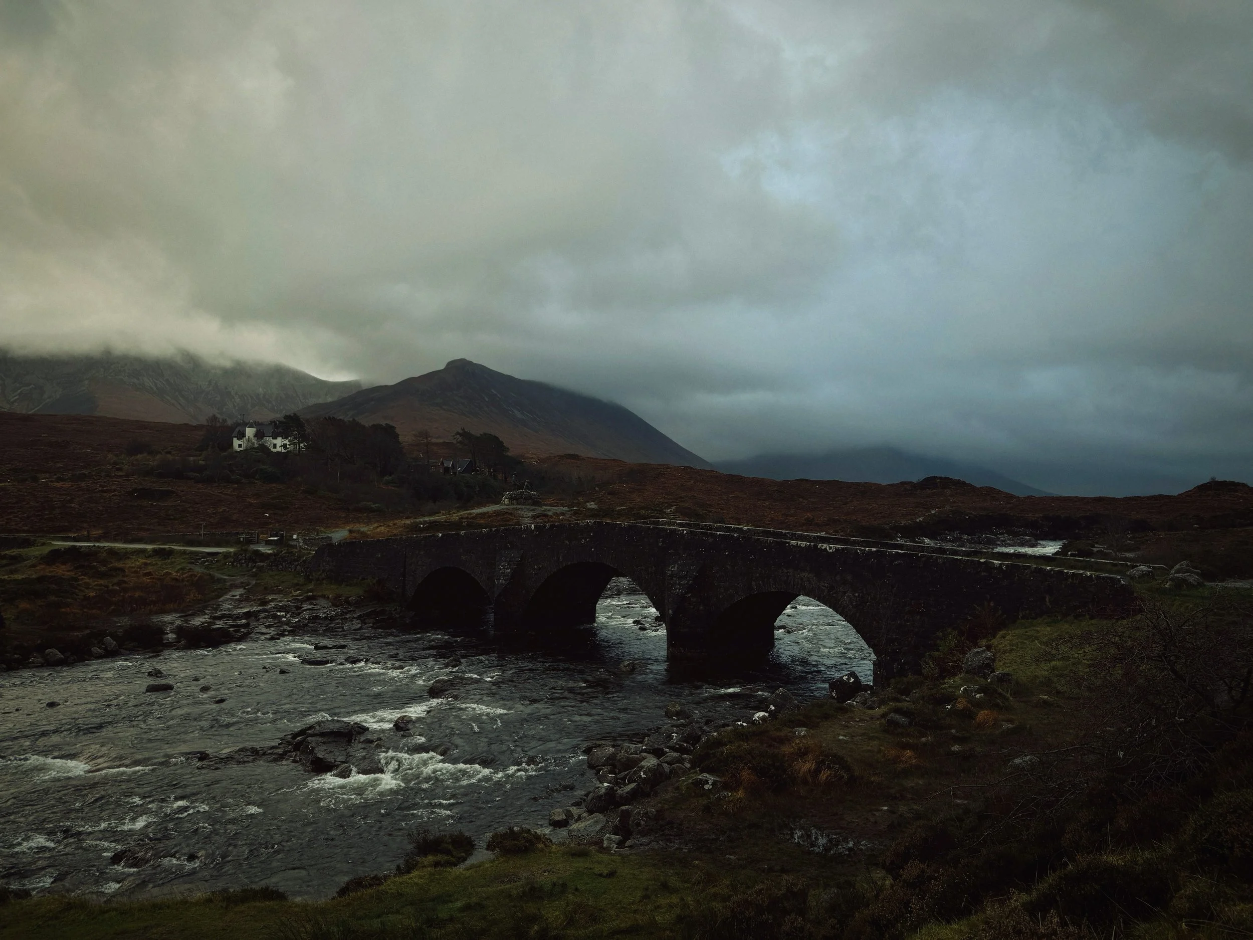 A dark and cloudy overcast landscape featuring a stone arch bridge over a rocky river, with hills and mountains in the background, and a few white houses on the hillside.