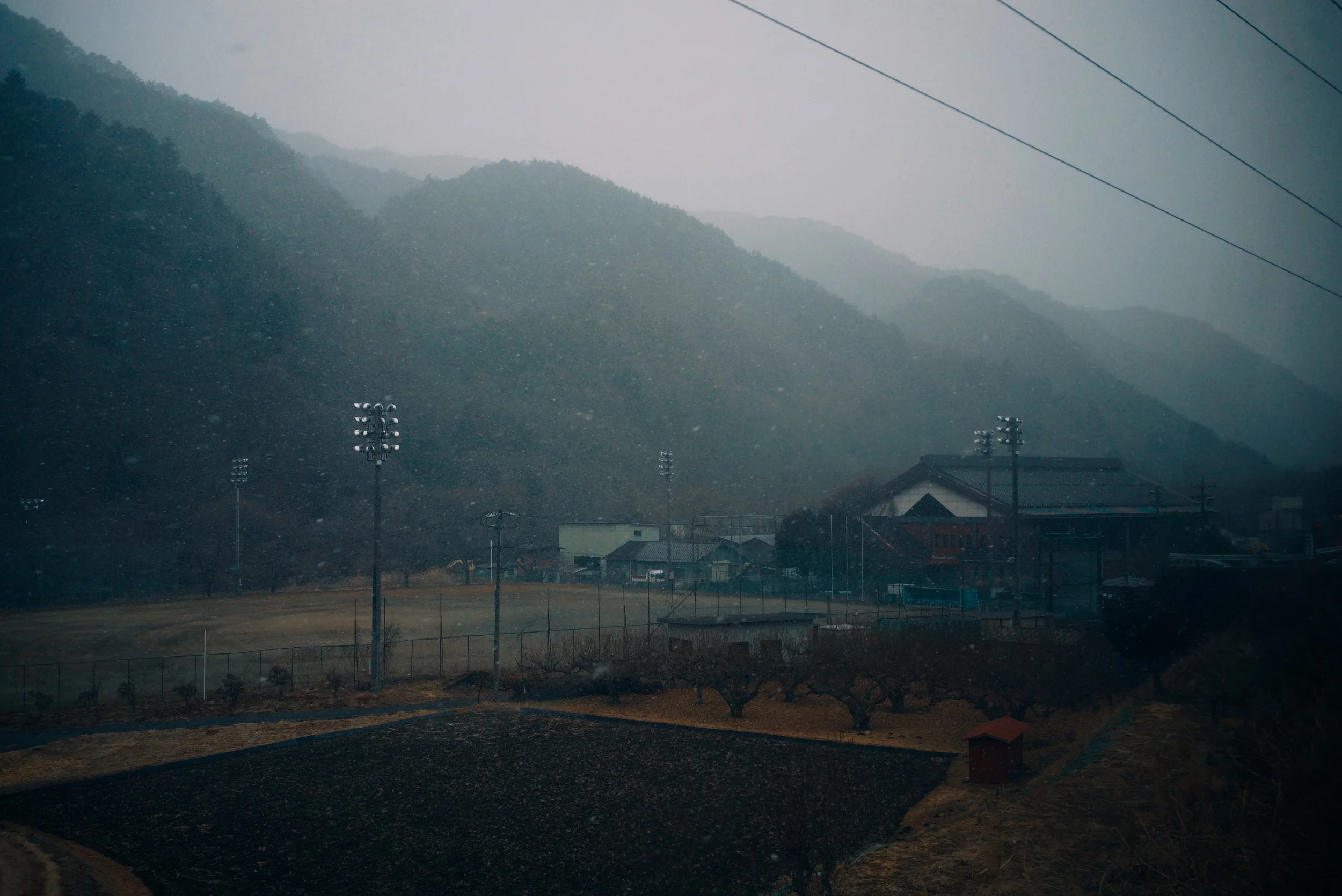 Rainy day in a rural village with mountains in the background, power lines overhead, and a sports field with tall floodlights in the foreground.