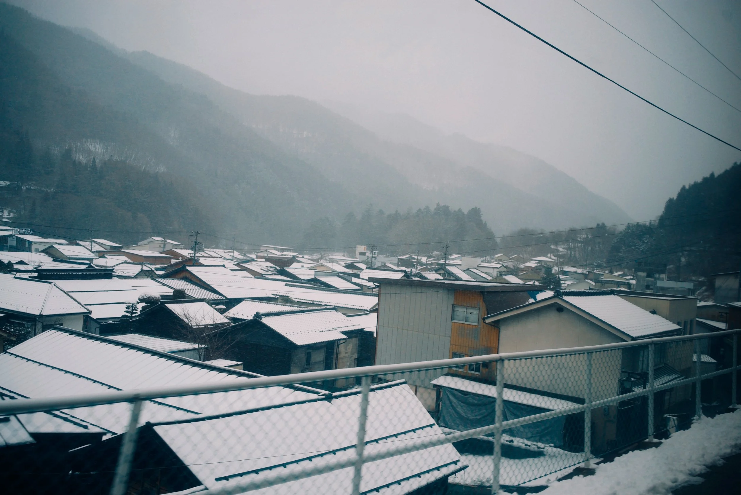 Snow-covered rooftops in a mountain town with misty mountains in the background and power lines overhead.