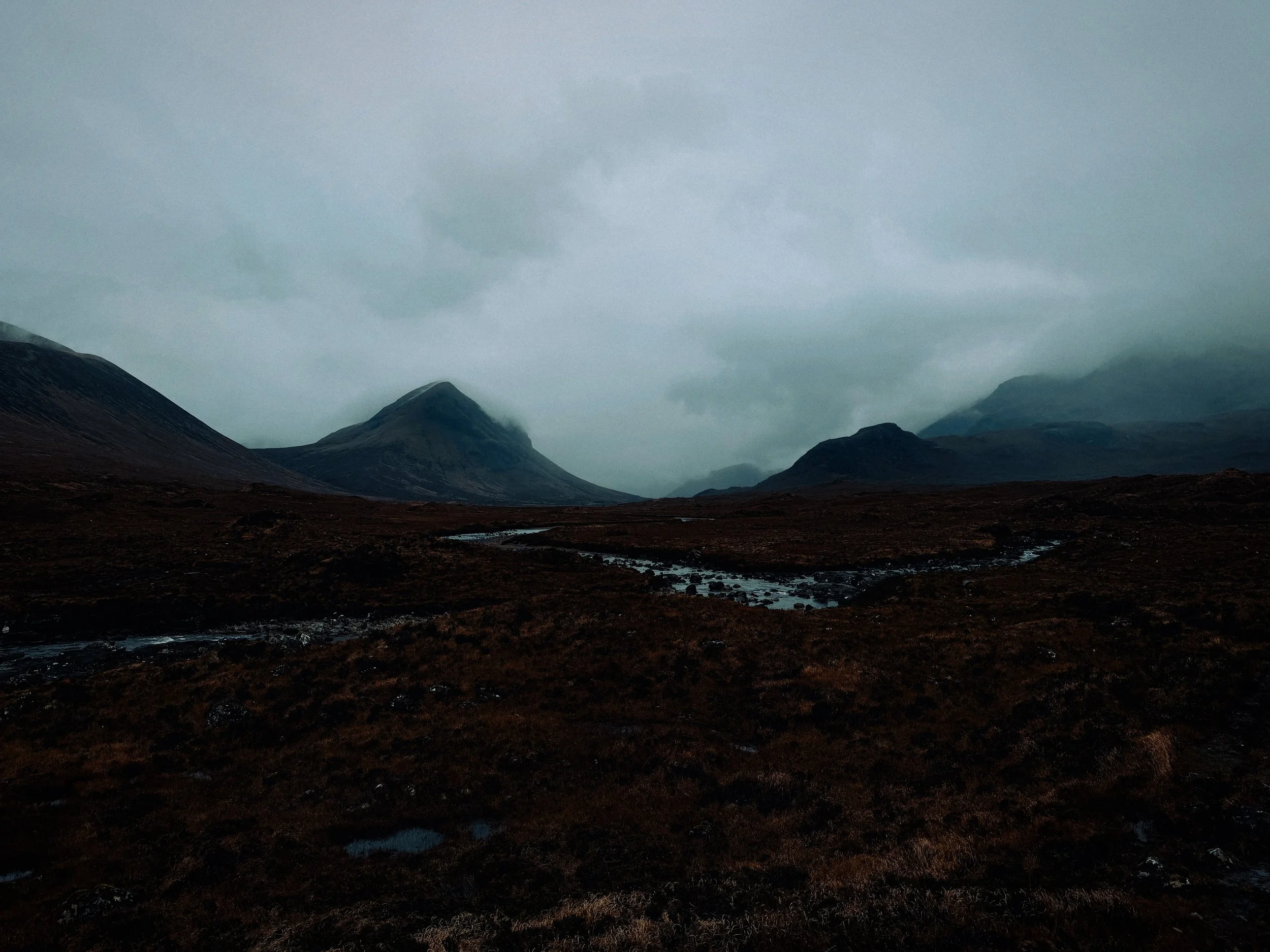 A dark, overcast landscape featuring mountains in the distance, a winding stream, and a moody sky.