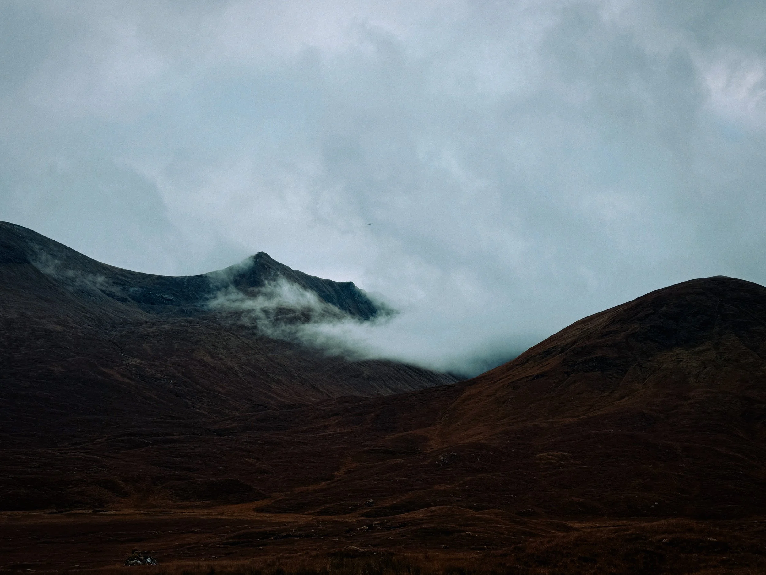 A mountain landscape with dark brown and gray rolling hills under a cloudy sky, with mist or low clouds around the mountain peaks.