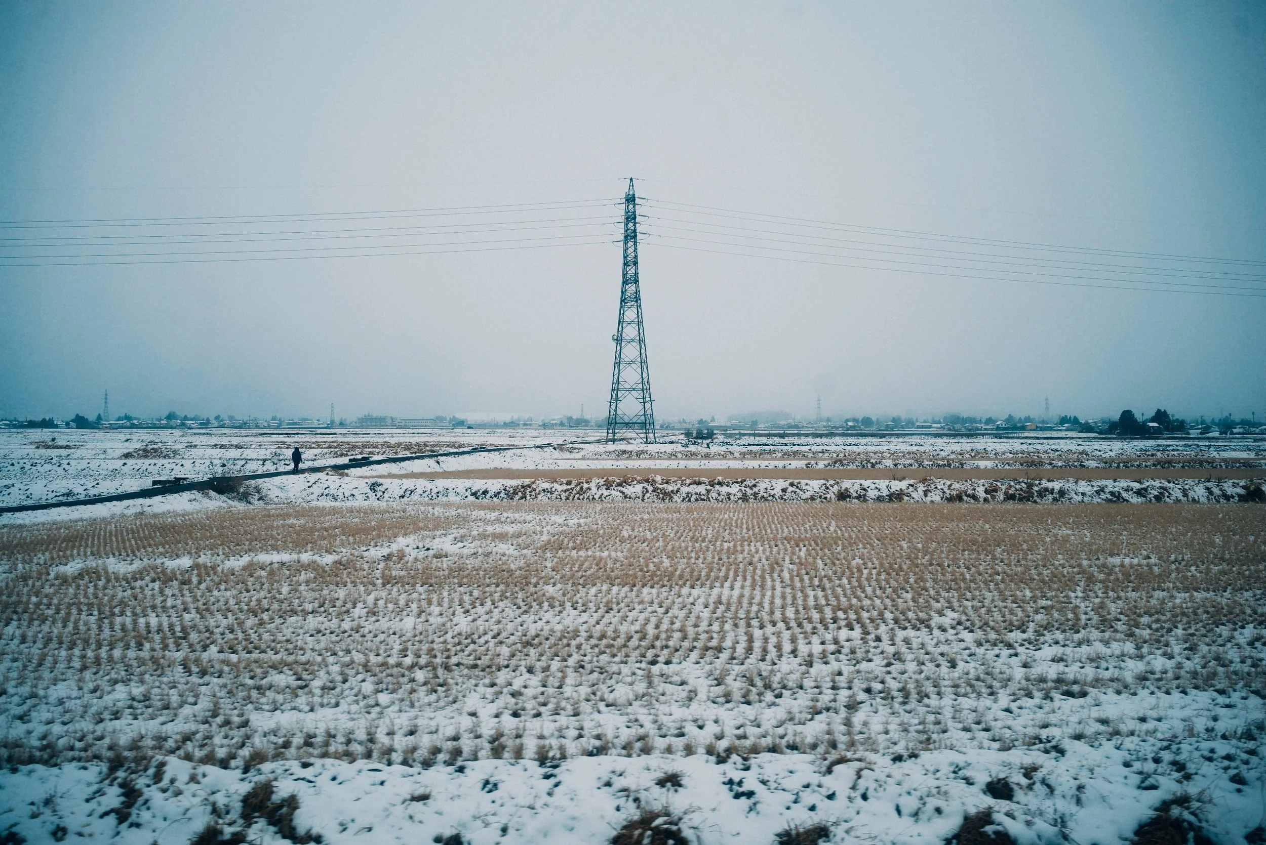 Snow-covered agricultural field with a person walking in distance and a tall power line tower in the middle, under a cloudy sky.