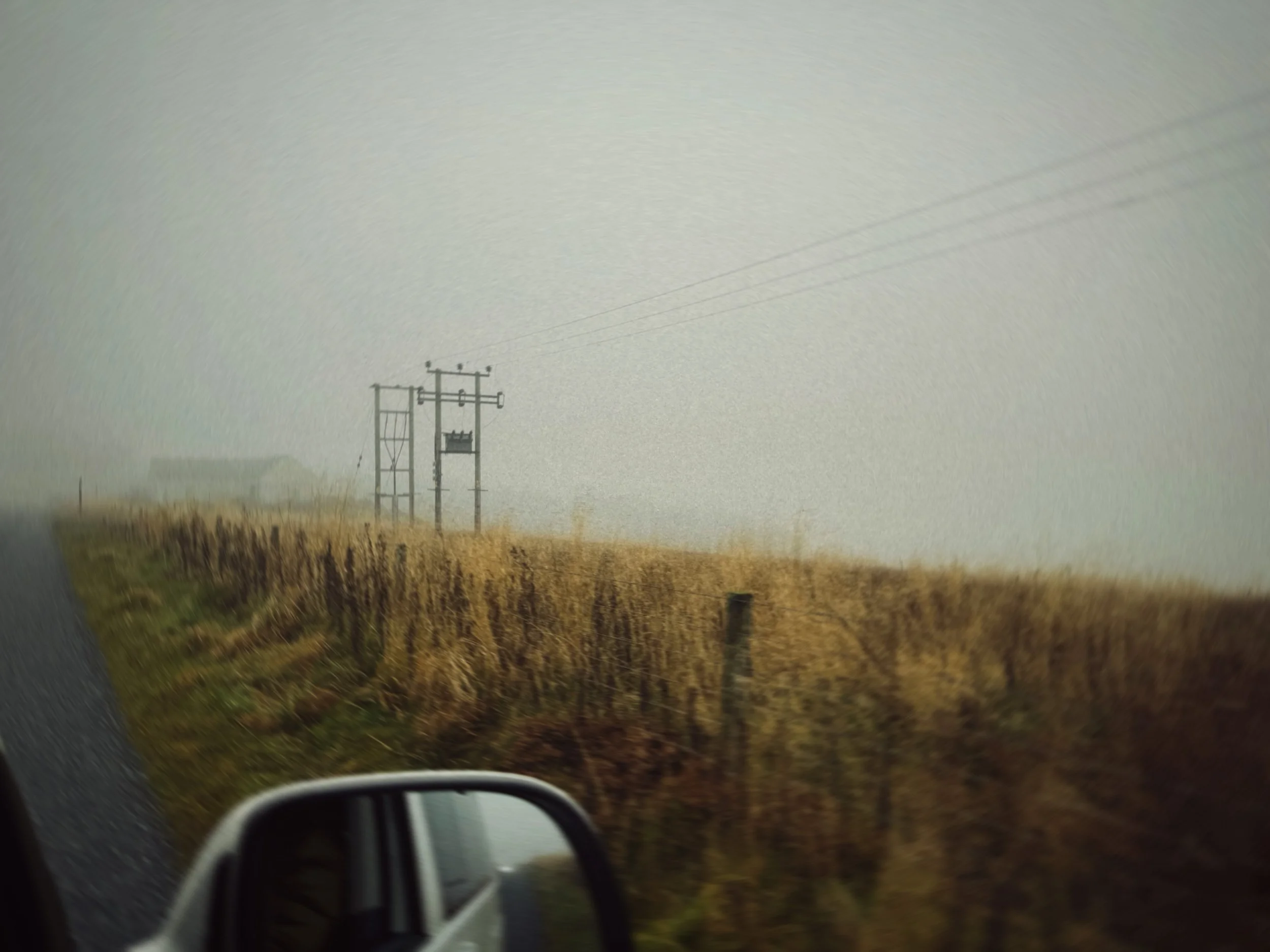 A foggy rural landscape with electrical poles and power lines along a field, with a side mirror of a vehicle visible in the foreground.