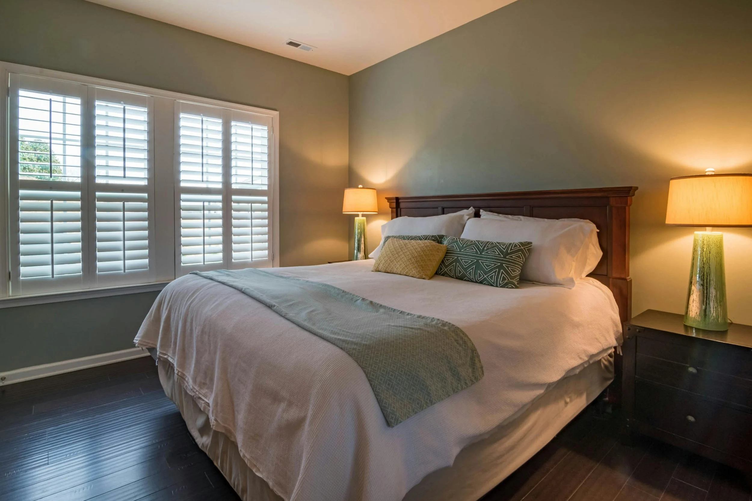 A bedroom with a bed, wooden headboard, green lamps, and windows with white plantation shutters.