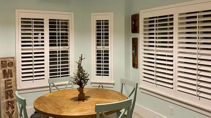 A dining room with light green walls, white plantation shutters on three windows, a round wooden table with four chairs, and a small decorative Christmas tree on the table.